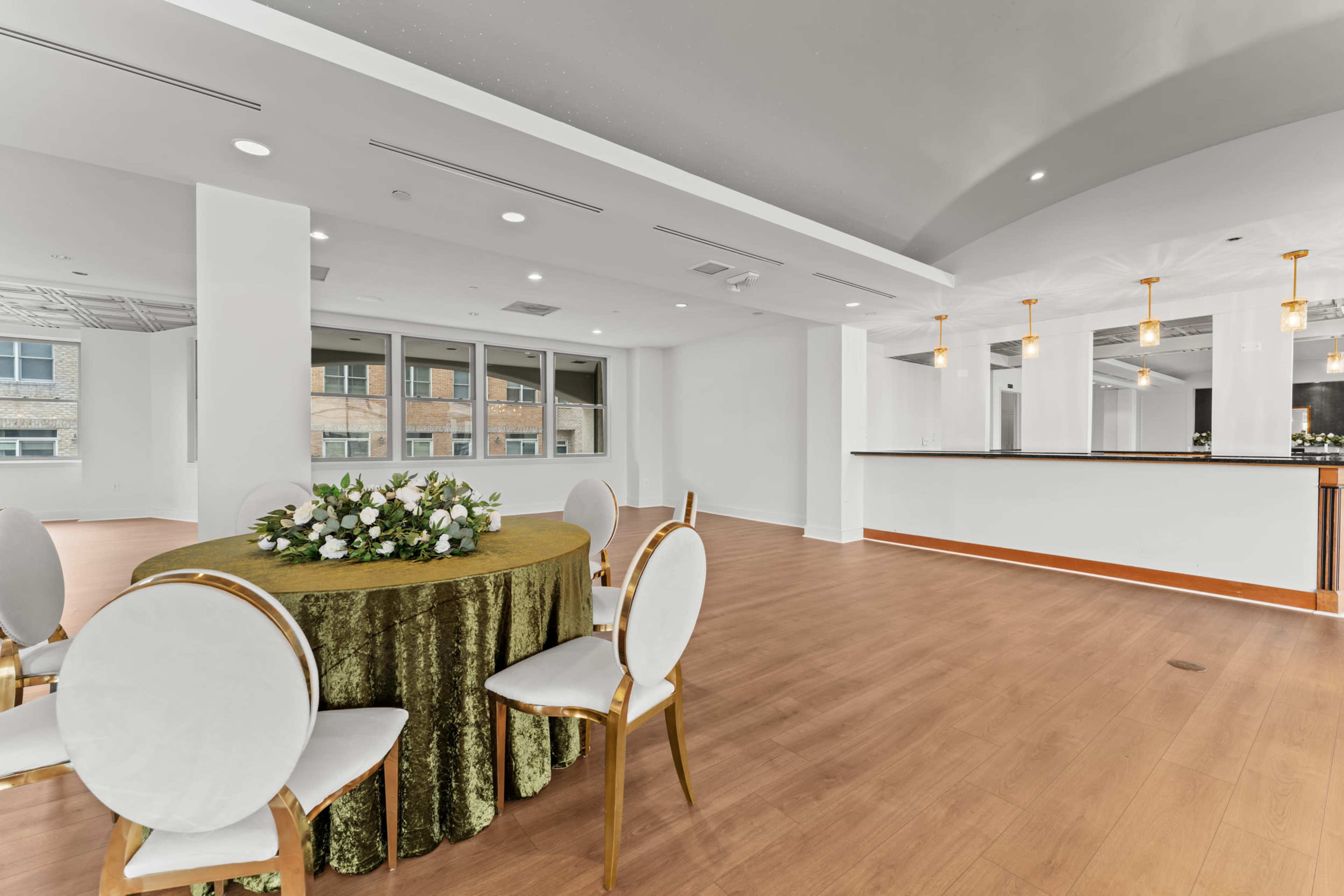 A round table with a green tablecloth and floral arrangement is set in a spacious, well-lit room featuring a bar area and large windows.