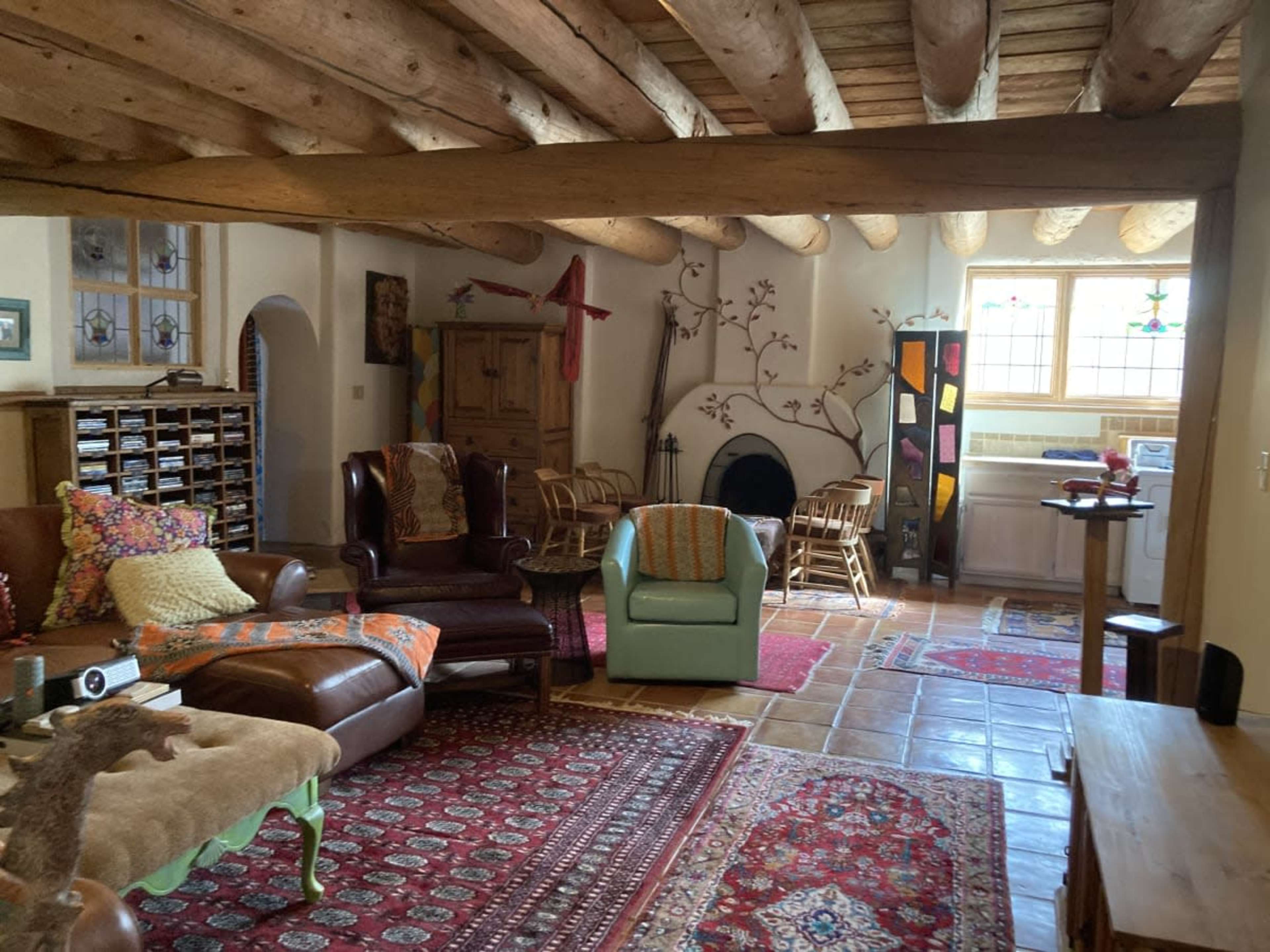 The room features wooden beams overhead, a fireplace, and a mix of seating including a leather couch and light blue armchair, surrounded by patterned rugs and various decorative elements.