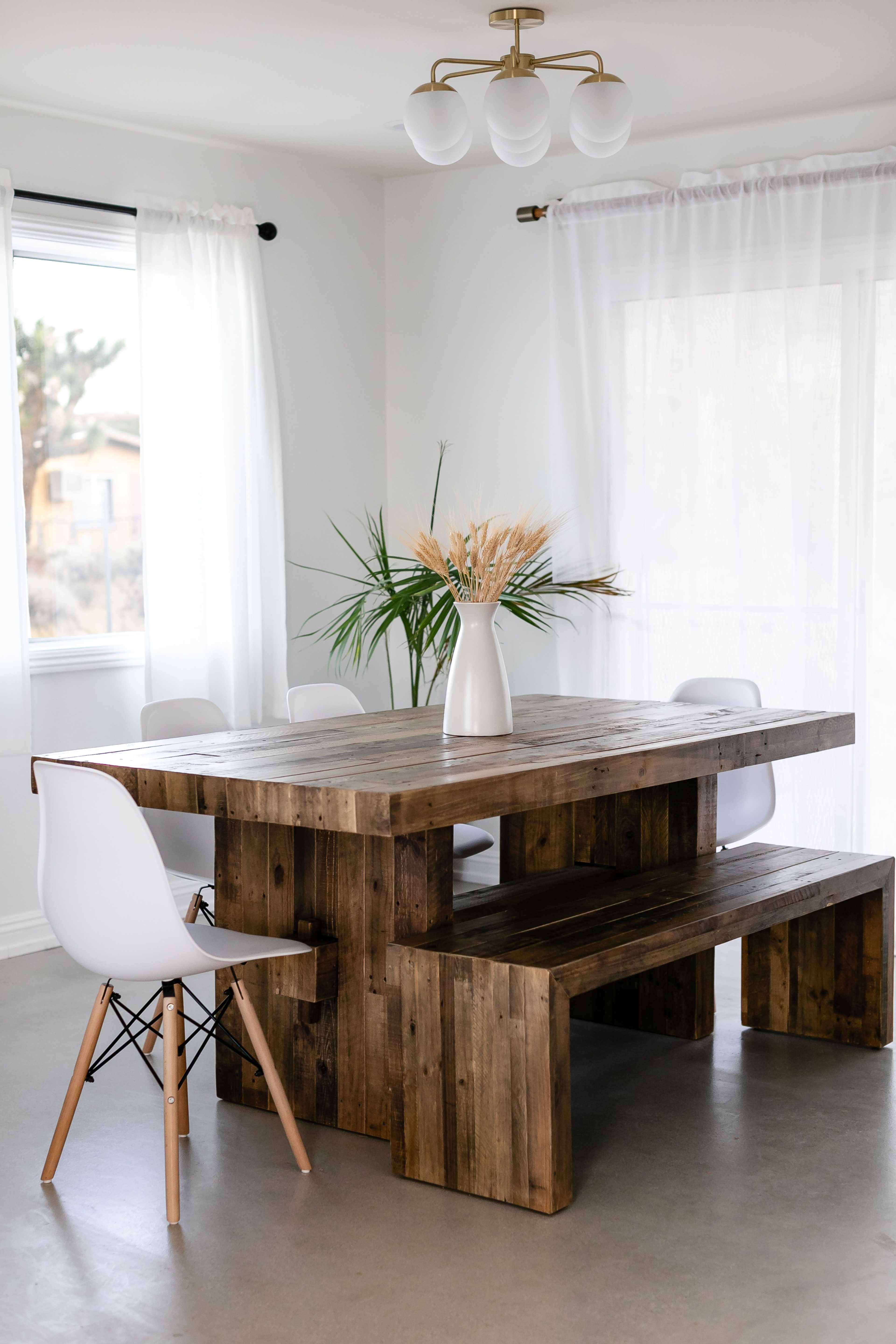 A wooden dining table with benches and white chairs, surrounded by large windows with sheer curtains.