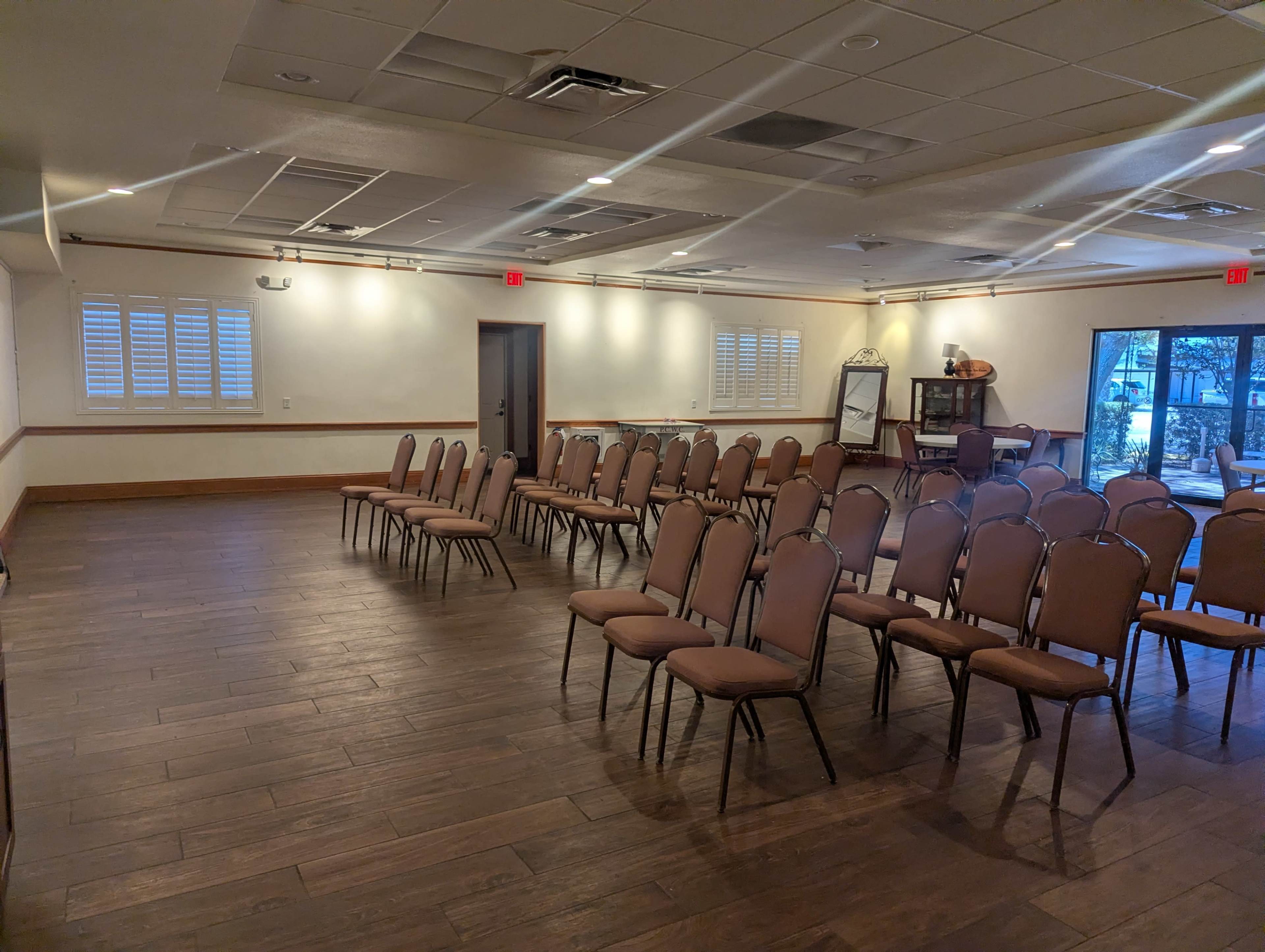 The image shows a room set up for an event, featuring several rows of empty chairs arranged on a wooden floor.
