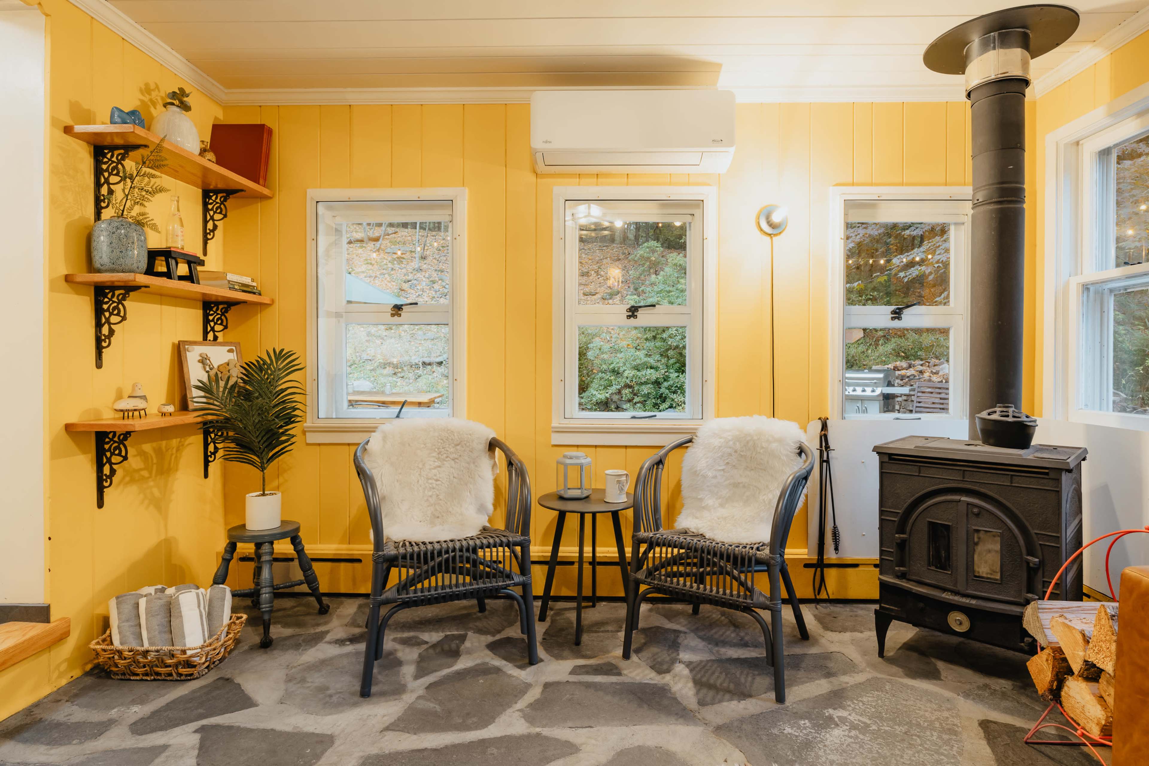 The scene features a cozy room with yellow walls, two black wicker chairs with white throws, a small circular table between them, a wood stove in the corner, and shelves displaying various decorative items.