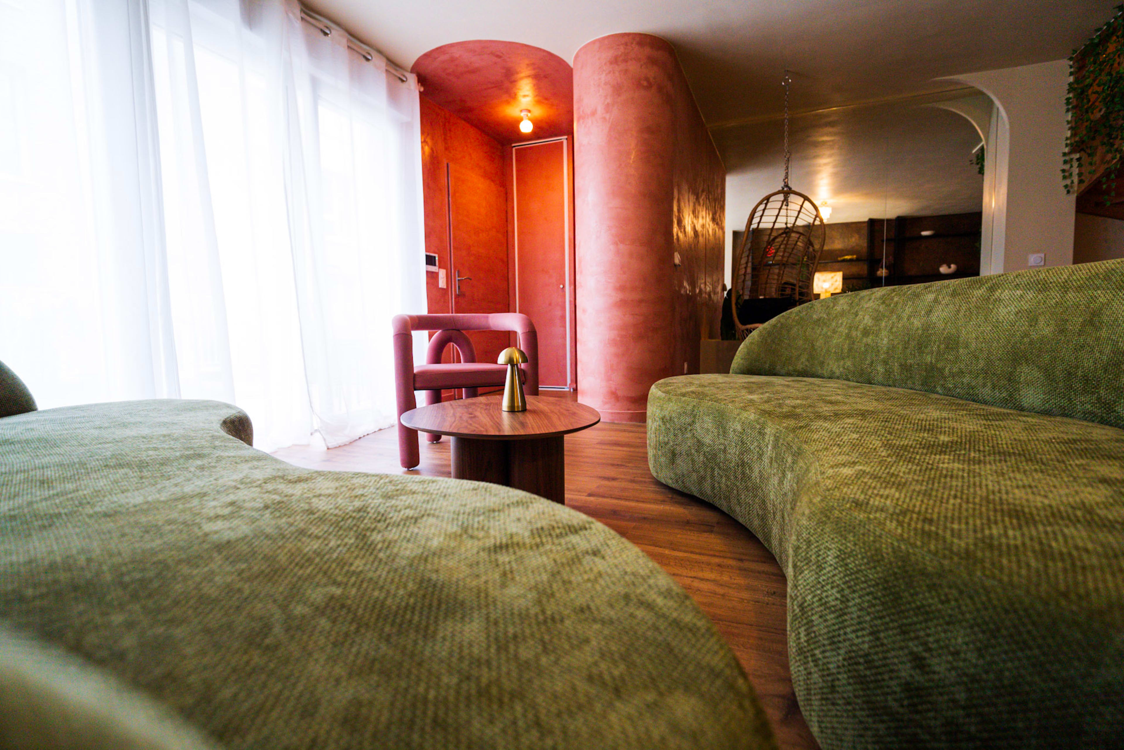 The image shows a modern living space with curved green sofas, a round wooden coffee table, and a pink feature wall.
