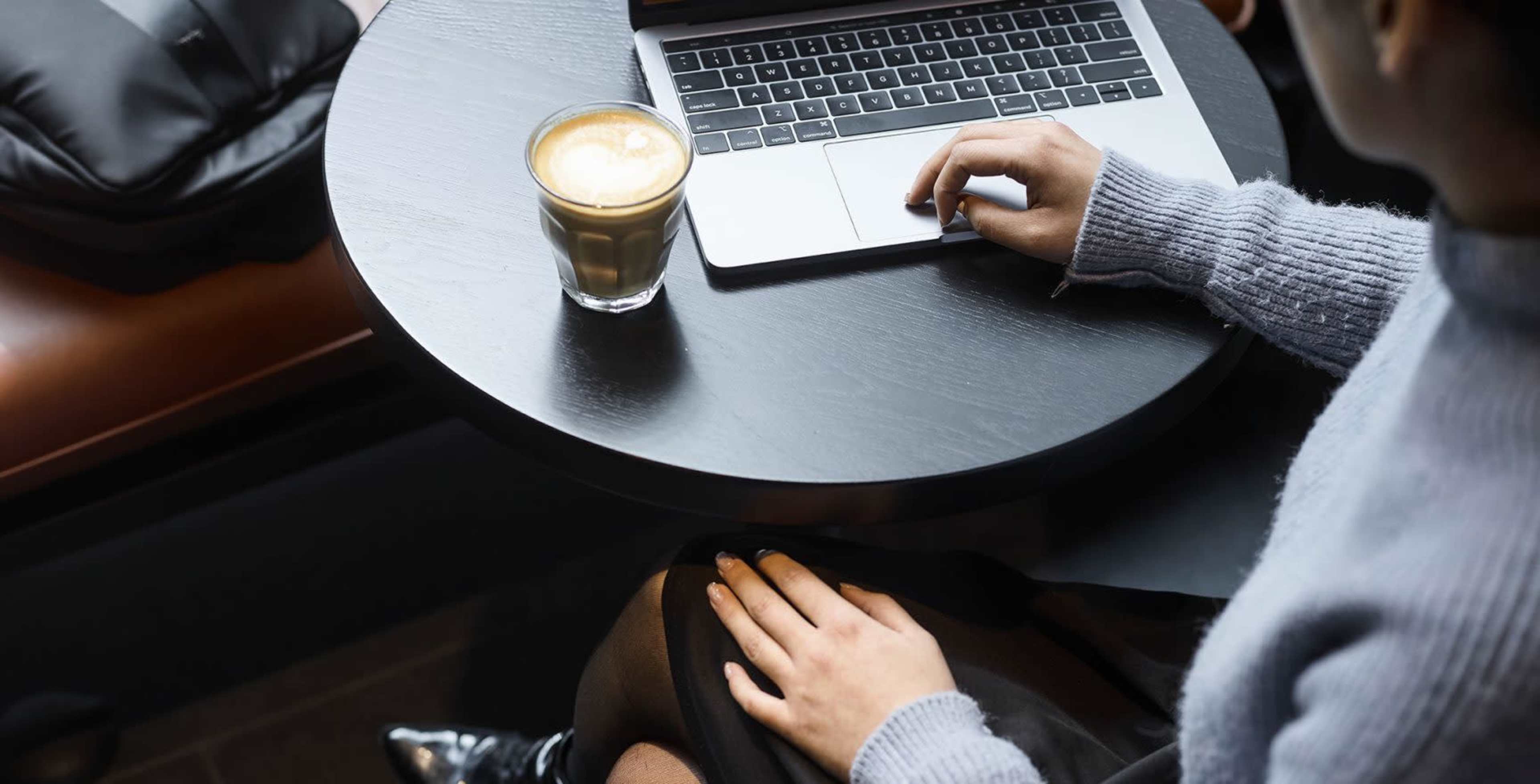 A person is seated at a black table, using a laptop while a glass of coffee sits beside them.