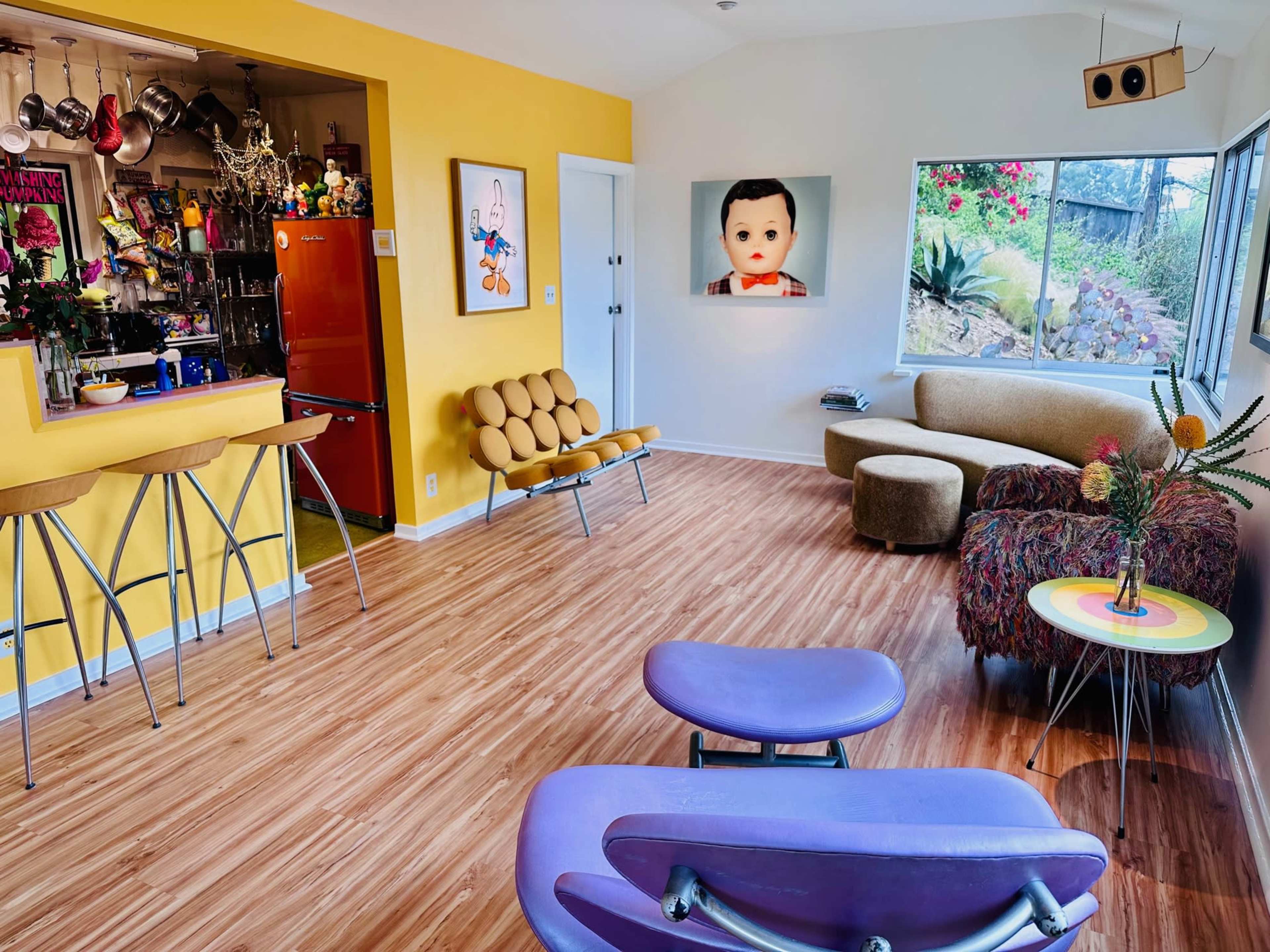 The image shows a brightly colored living room decorated with modern furniture, featuring a yellow accent wall and large windows overlooking a garden.