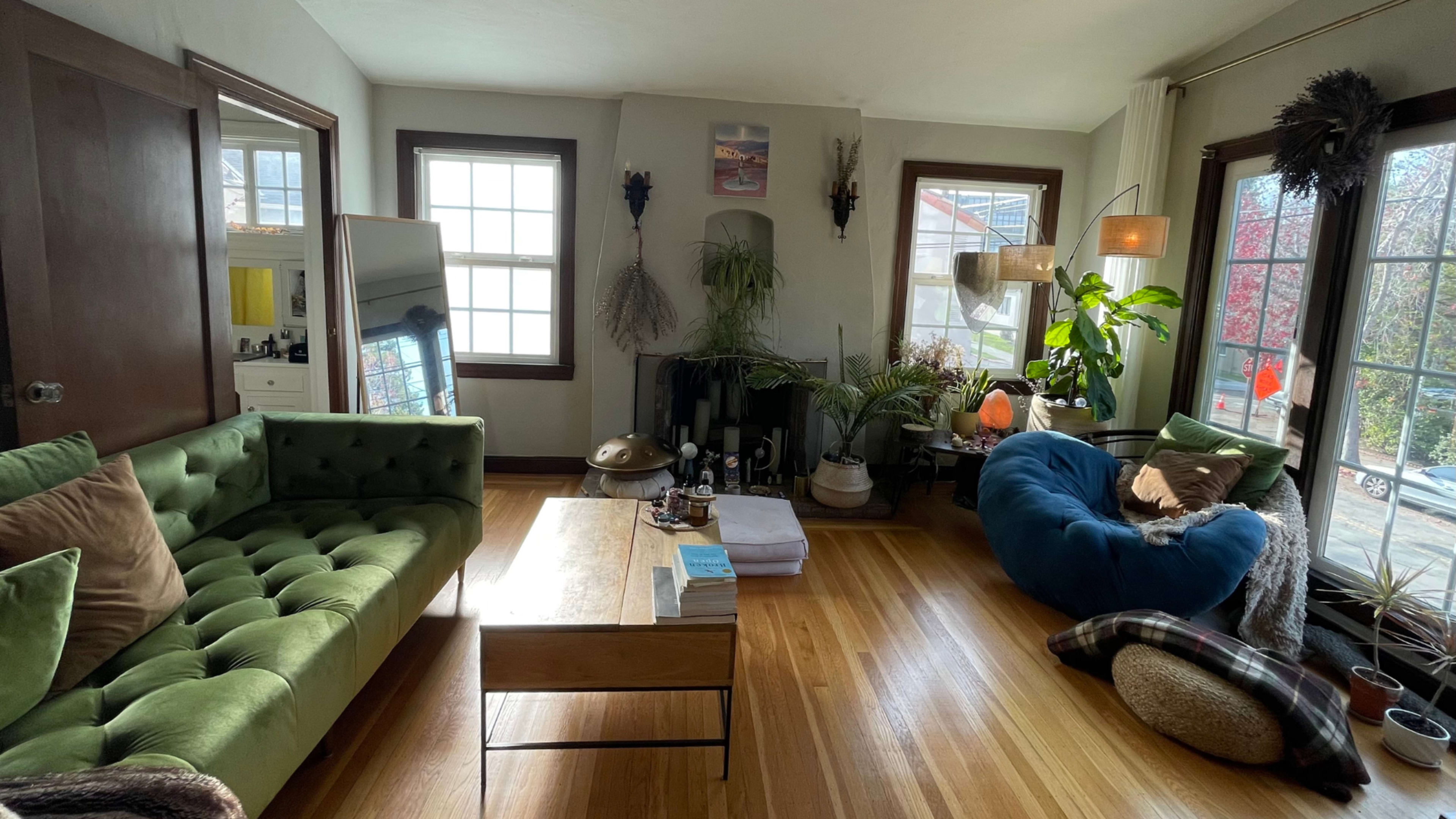 The living room features a green tufted sofa, a wooden coffee table, a beanbag chair, and large windows that provide natural light, along with various plants and decor.