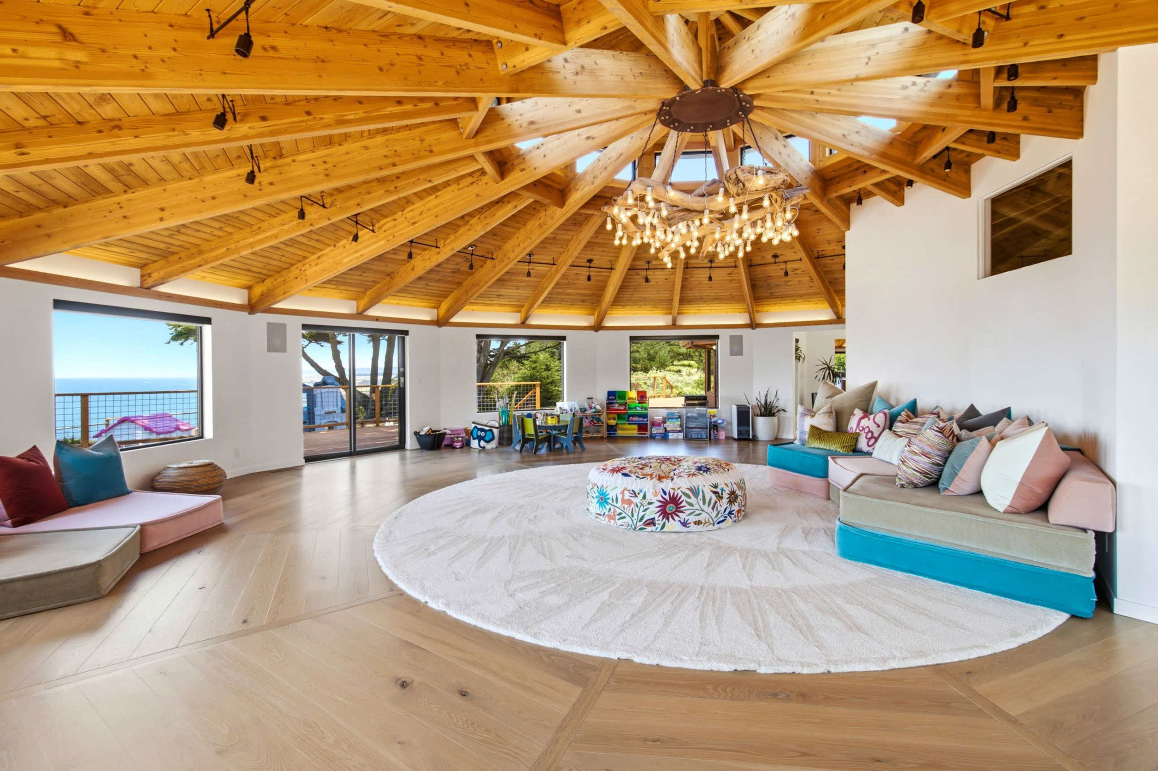 The image depicts a spacious circular room with a wooden ceiling featuring a chandelier, surrounded by large windows that offer a view of the ocean.