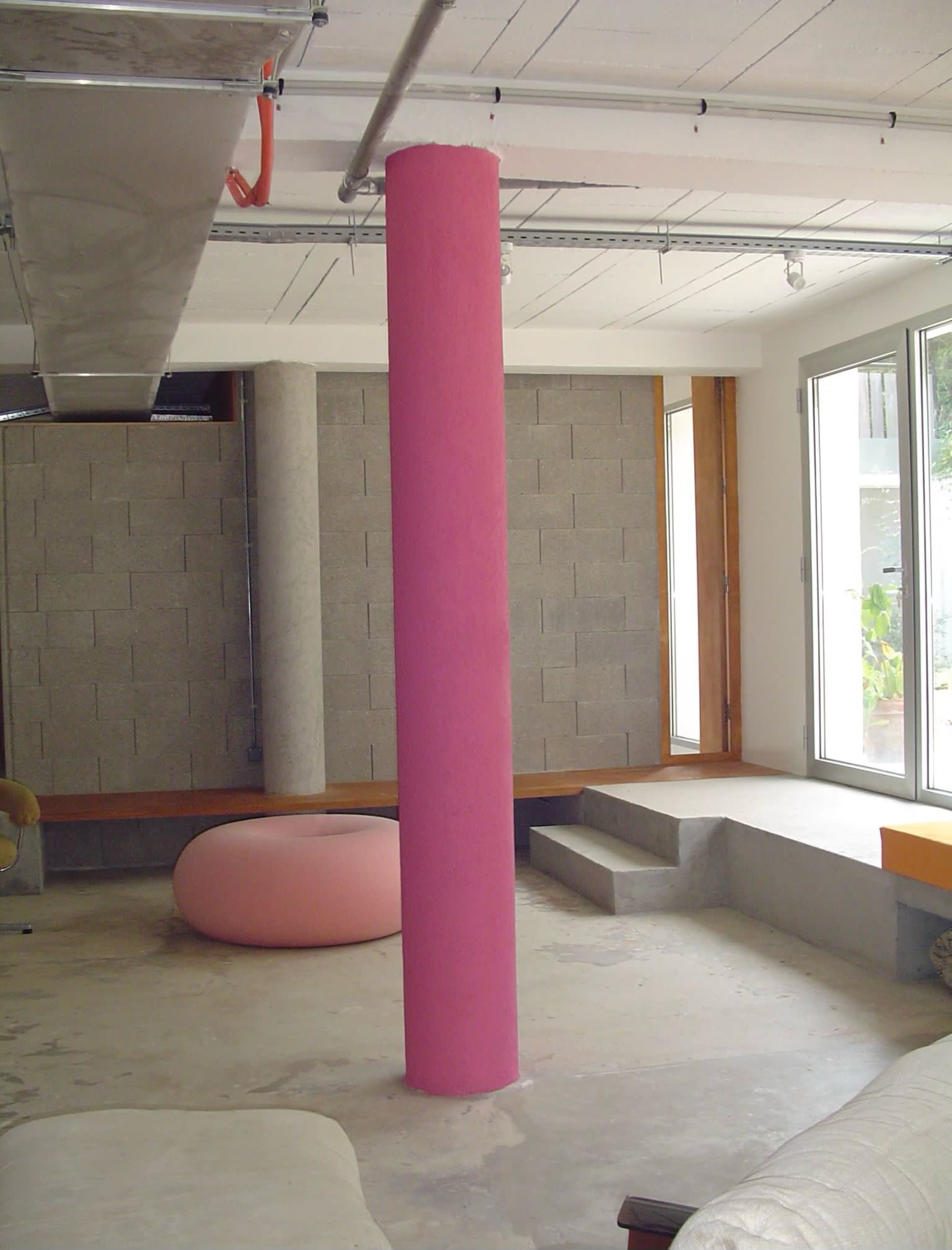 The image shows a modern interior space featuring a pink cylindrical pillar, concrete walls, and a stepped concrete area leading to large windows.