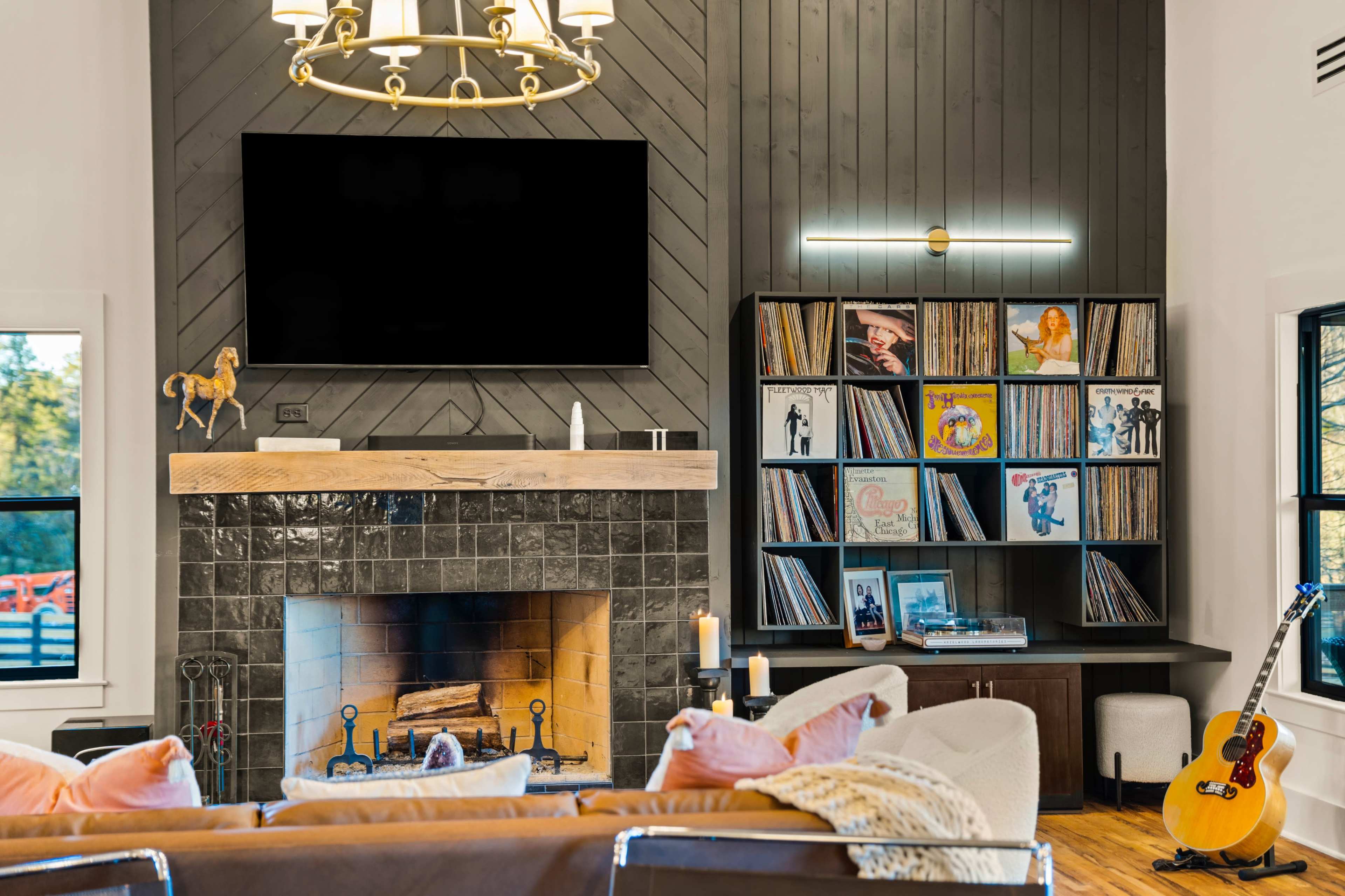 A living room features a fireplace, a large flat-screen TV above the mantel, and a wall-mounted shelf displaying a collection of vinyl records.
