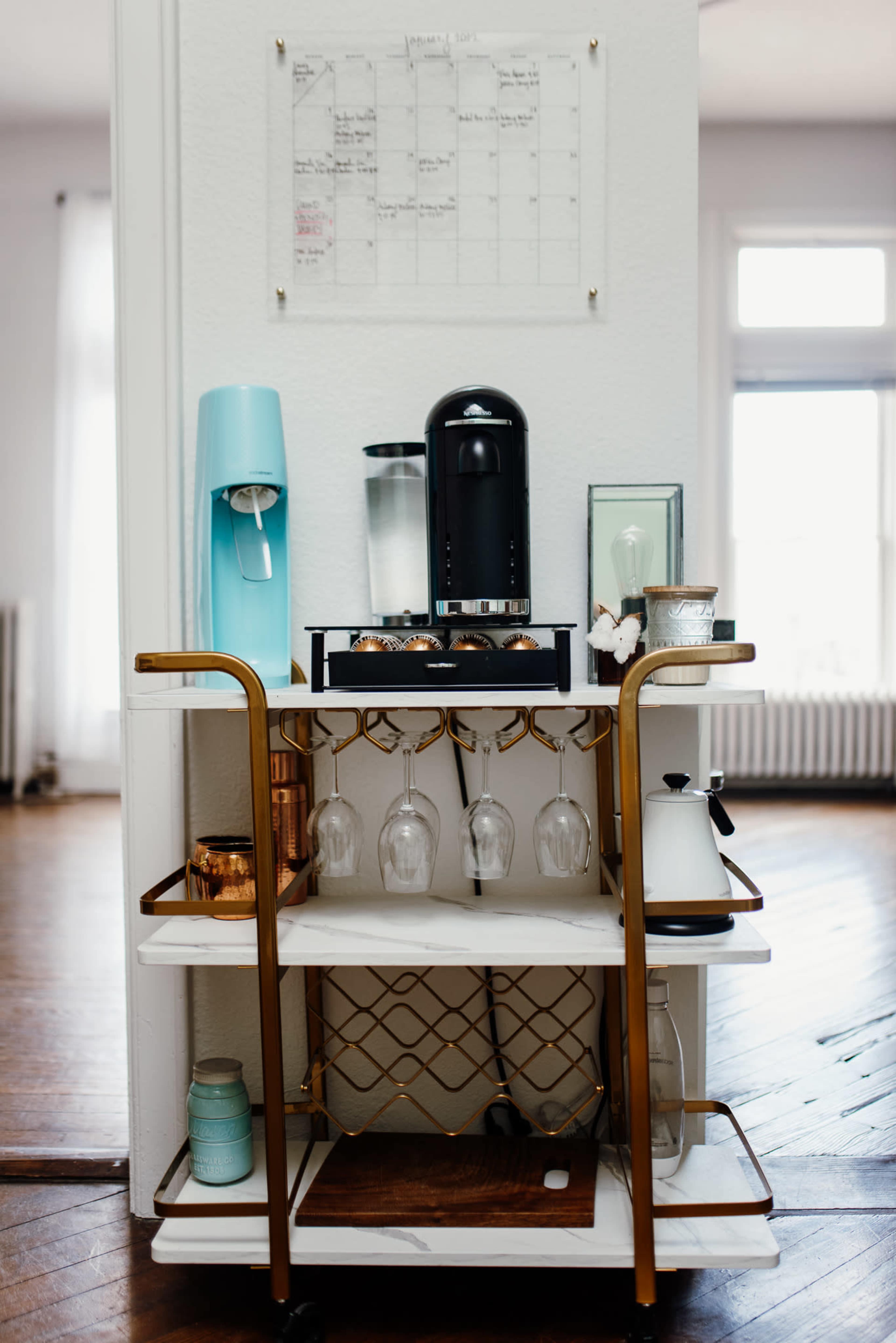 A bar cart featuring a coffee machine, glassware, and a water dispenser is positioned against a wall with a calendar above it.