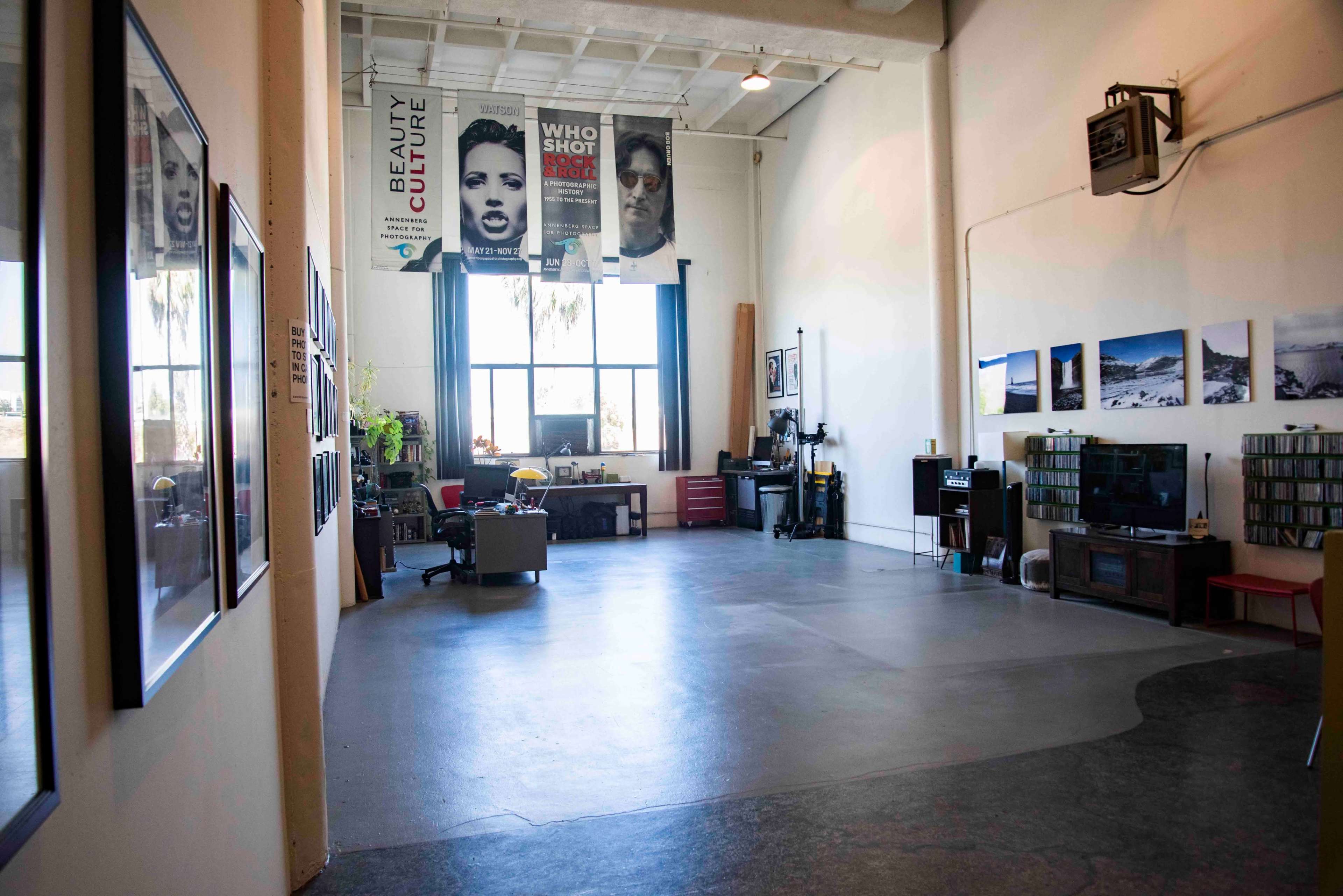 A spacious studio or gallery with posters on the walls, a large window letting in natural light, and various furnishings including a sofa and shelves.