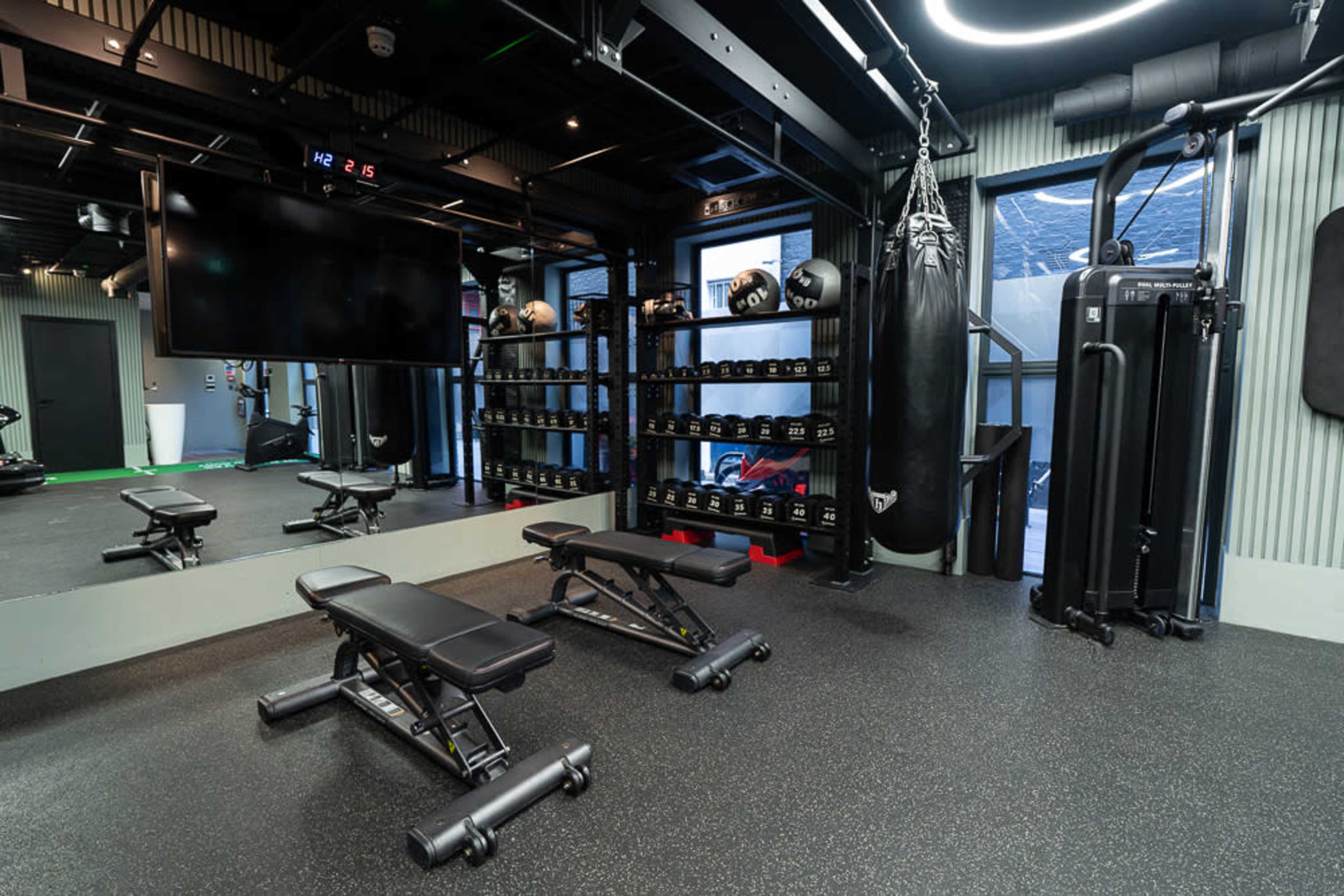 The image shows a modern gym interior featuring weight benches, dumbbells on shelves, a punching bag, and various exercise equipment.