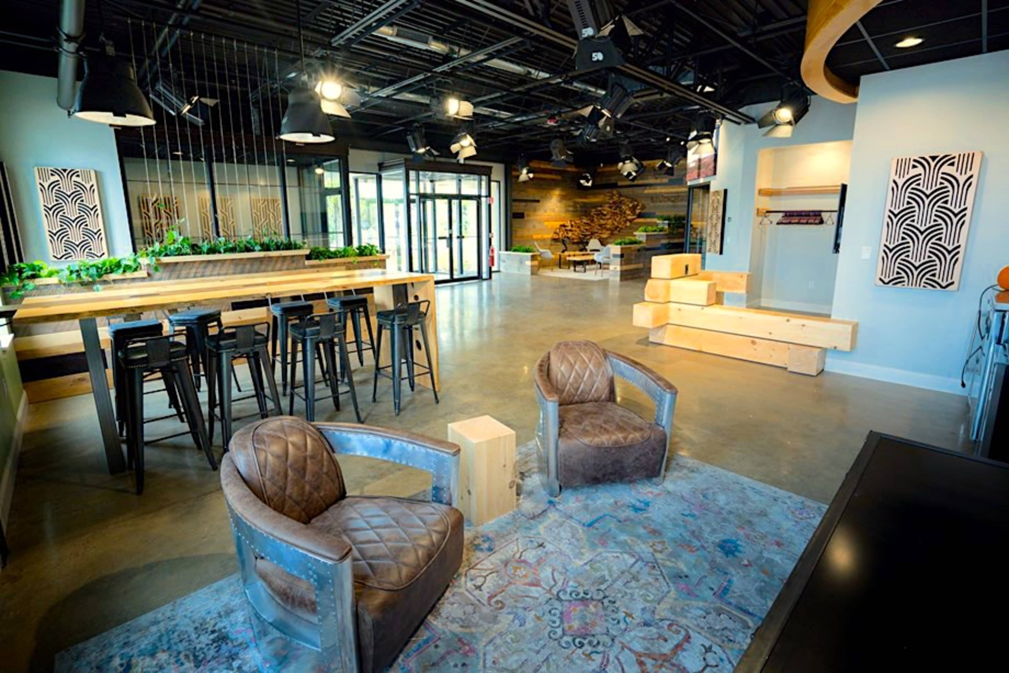 The image shows a modern open space with a seating area, bar-style tables, and decorative plants.