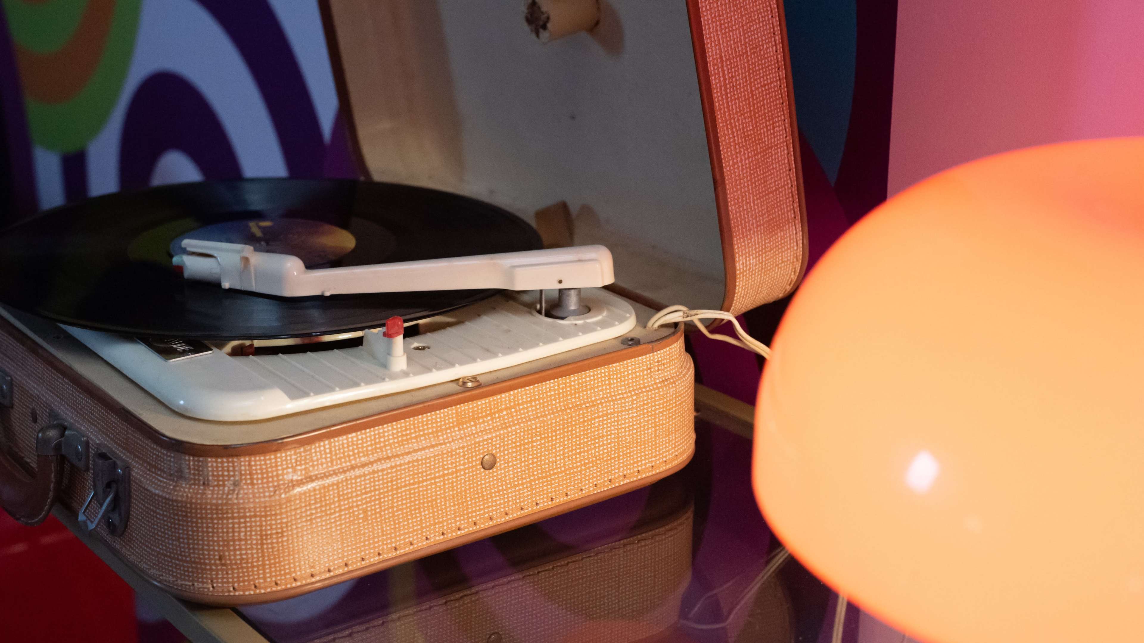 A vintage record player is positioned next to an orange lampshade on a colorful patterned background.