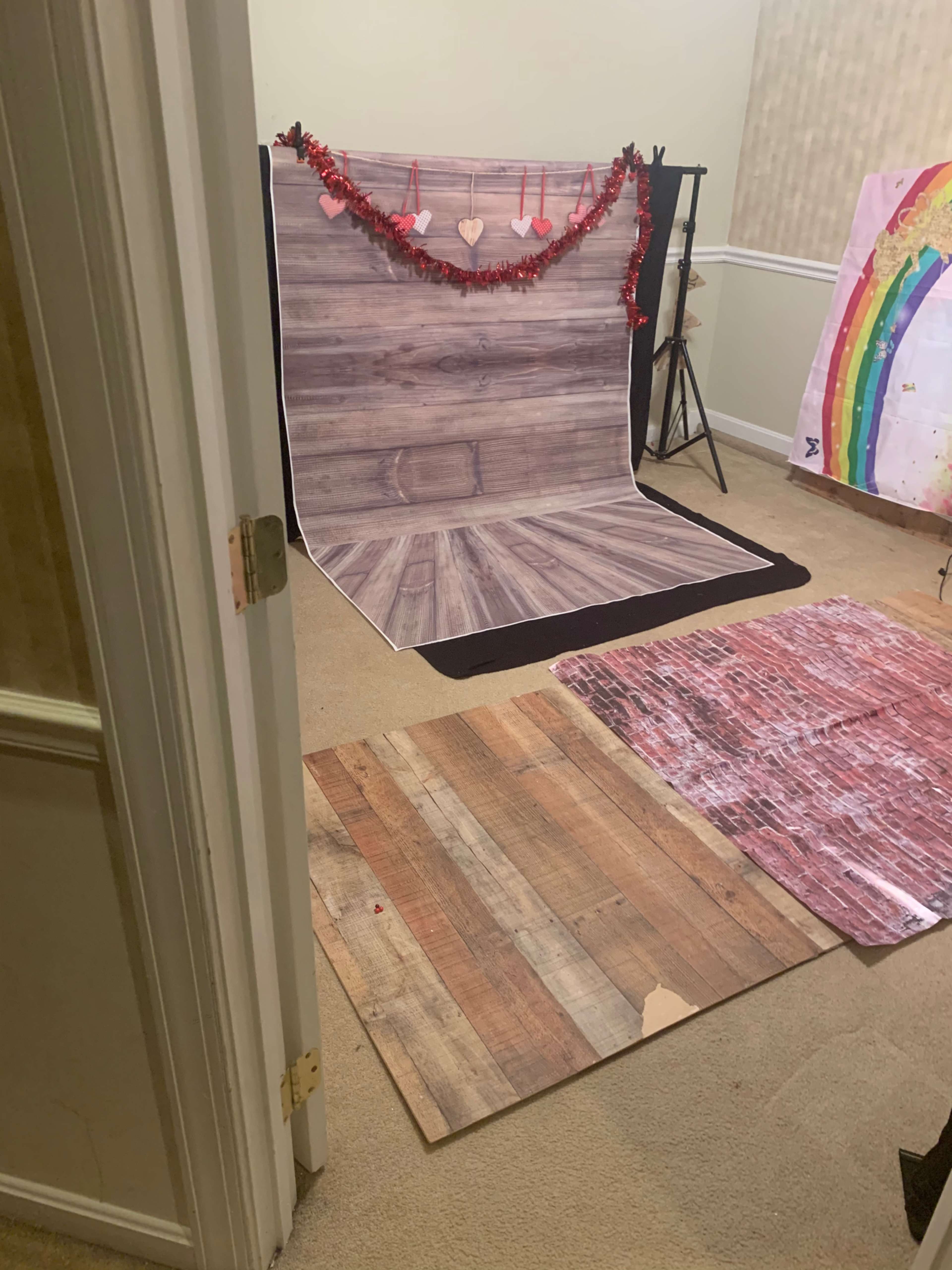 A photography setup with a wooden backdrop, decorative hearts, and various floor mats in a carpeted room.