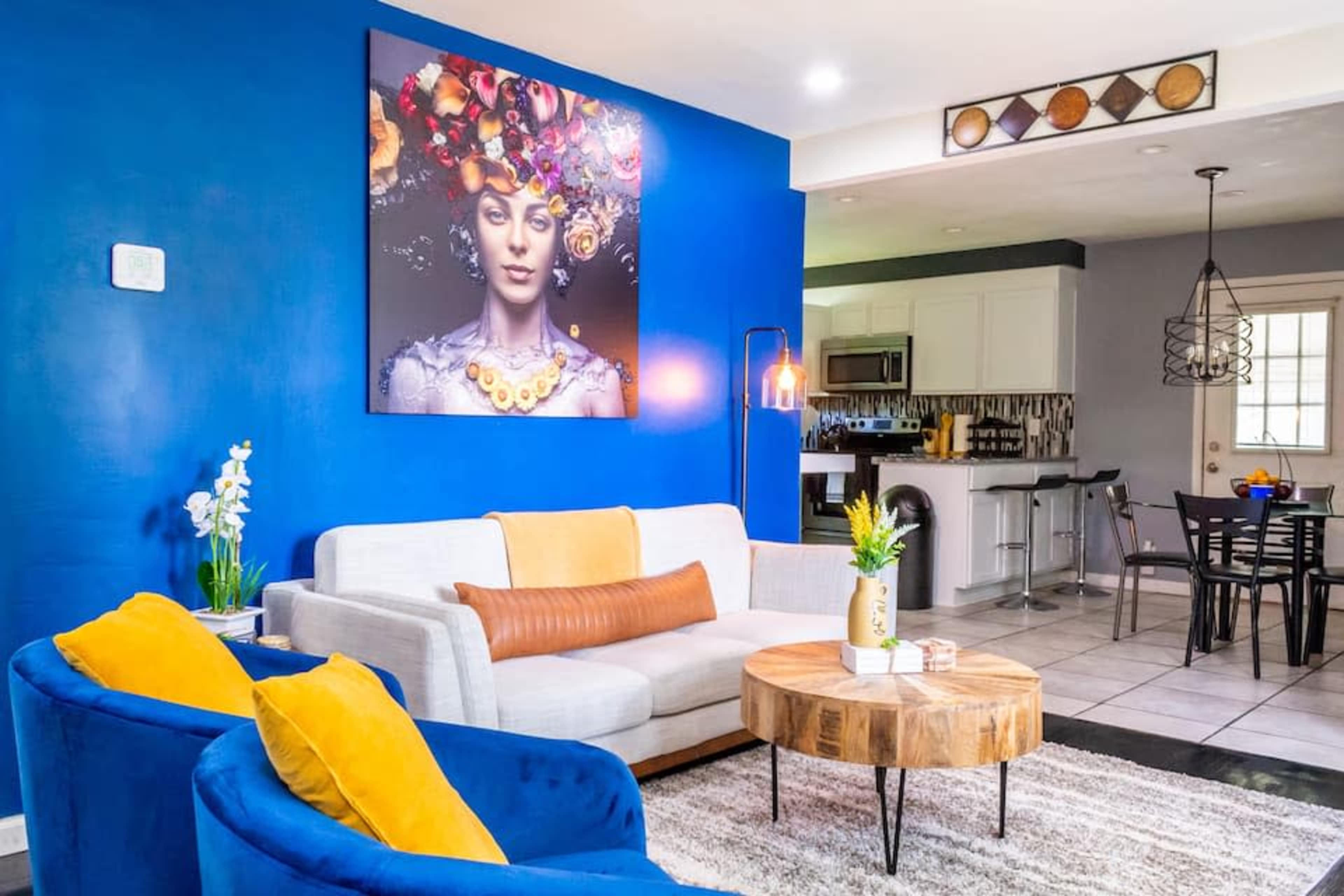 A modern living room with blue walls, a large artwork featuring a woman with flowers in her hair, a gray sofa, yellow accent chairs, and a round wooden coffee table.