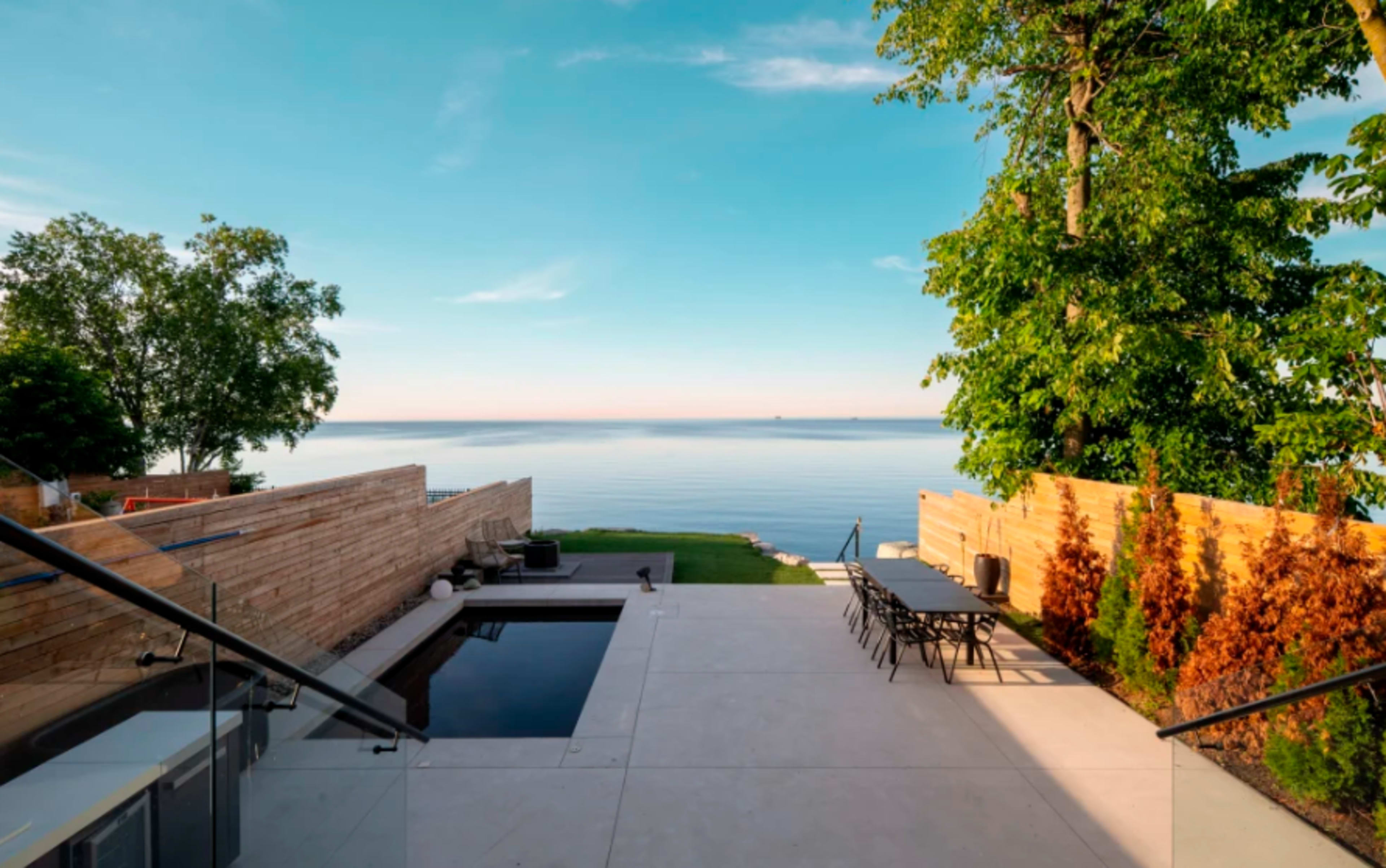 The scene features a modern patio overlooking a calm body of water, with a swimming pool and landscaped greenery.