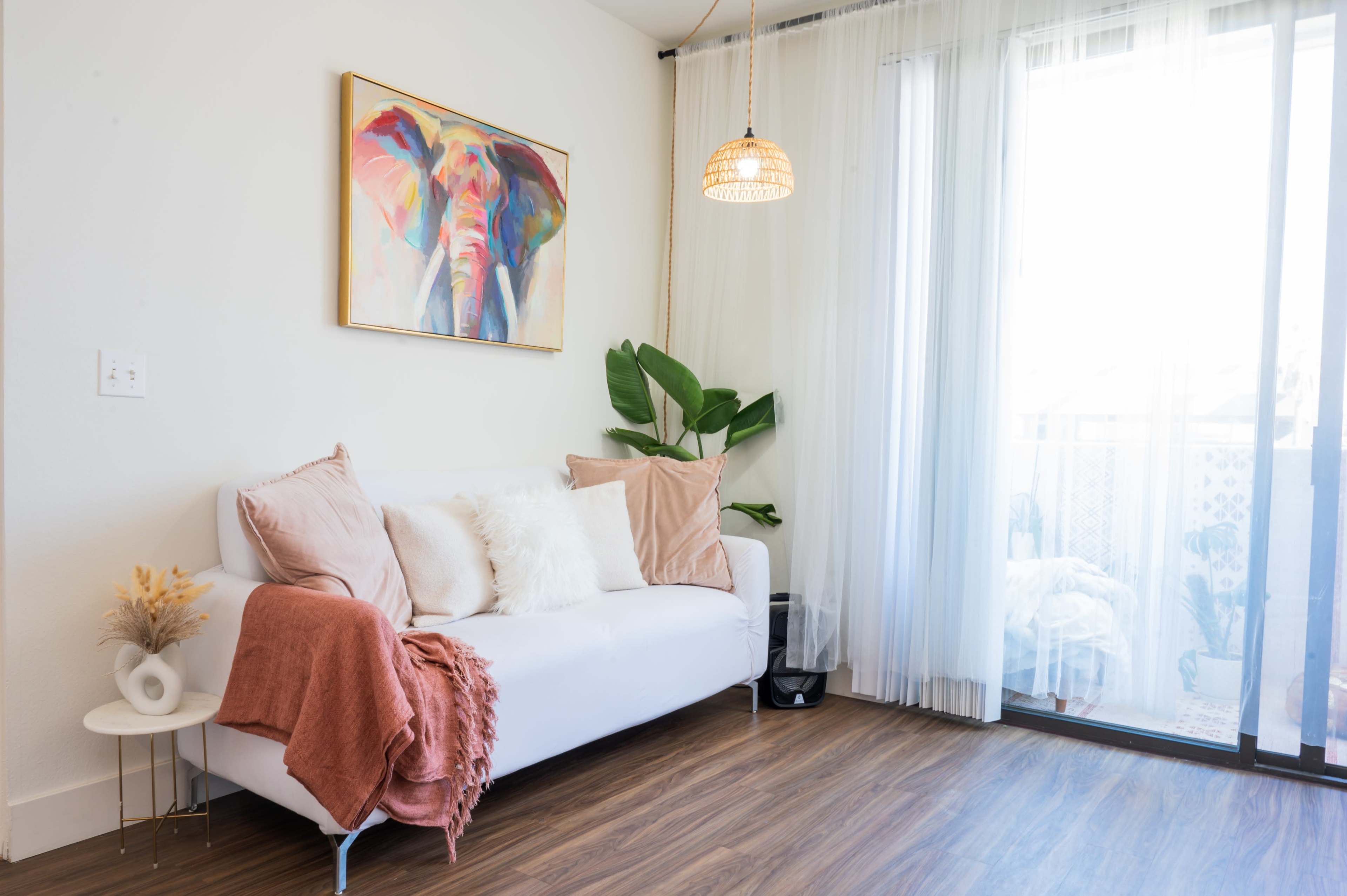 The living room features a white sofa adorned with soft cushions and a throw, positioned near a large window with sheer curtains and a colorful elephant painting on the wall.