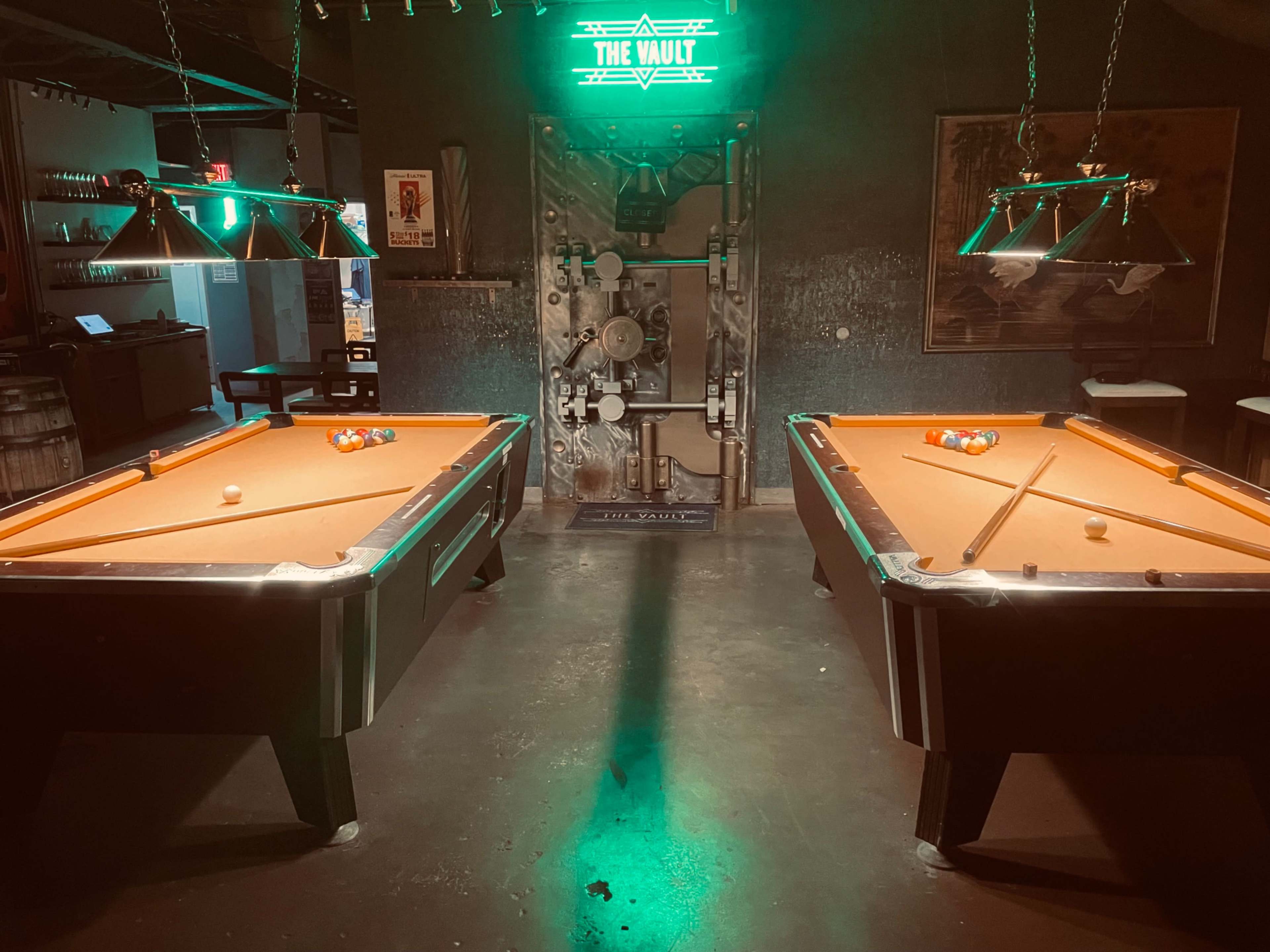 The image shows two pool tables with orange felt in a dimly lit room, illuminated by green neon lights and featuring a decorative vault door in the background.
