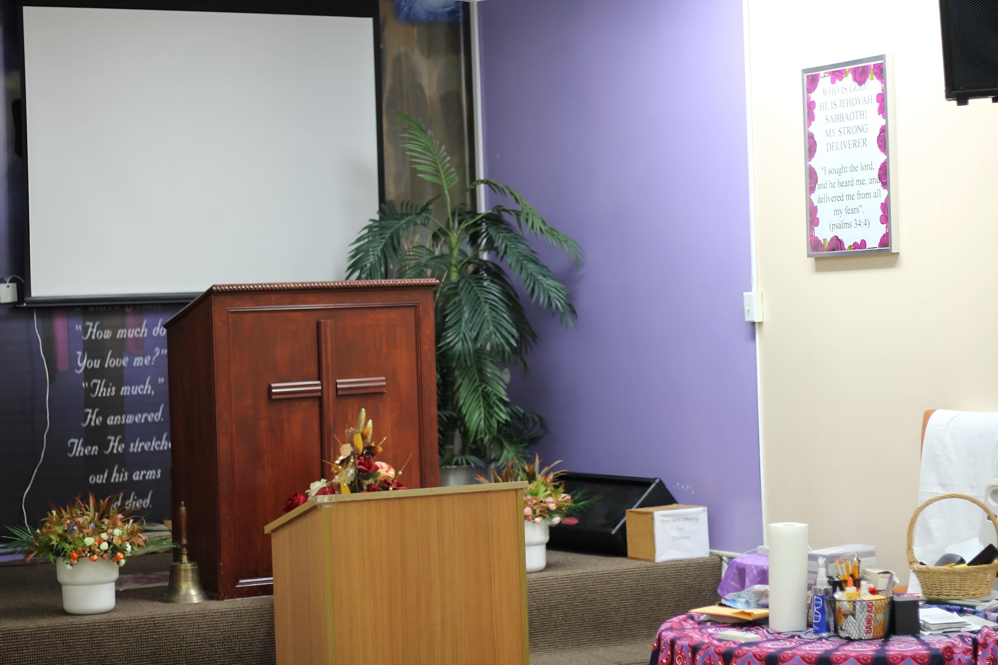 The image shows a small church setup featuring a wooden pulpit, a decorative table with items, and a large screen in the background.