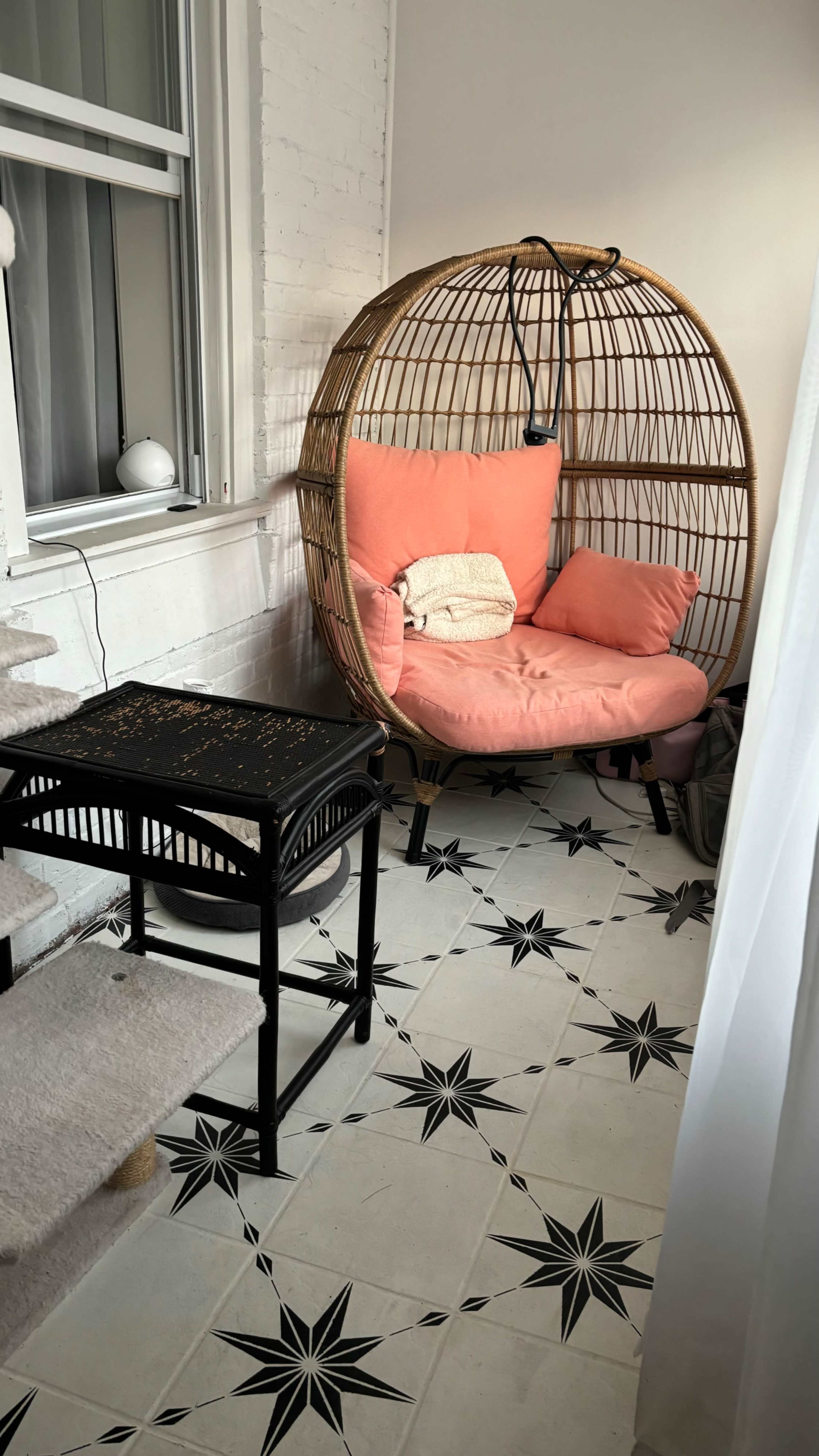 A rattan egg chair with a coral cushion sits next to a small black table on a star-patterned tiled floor in a well-lit corner.