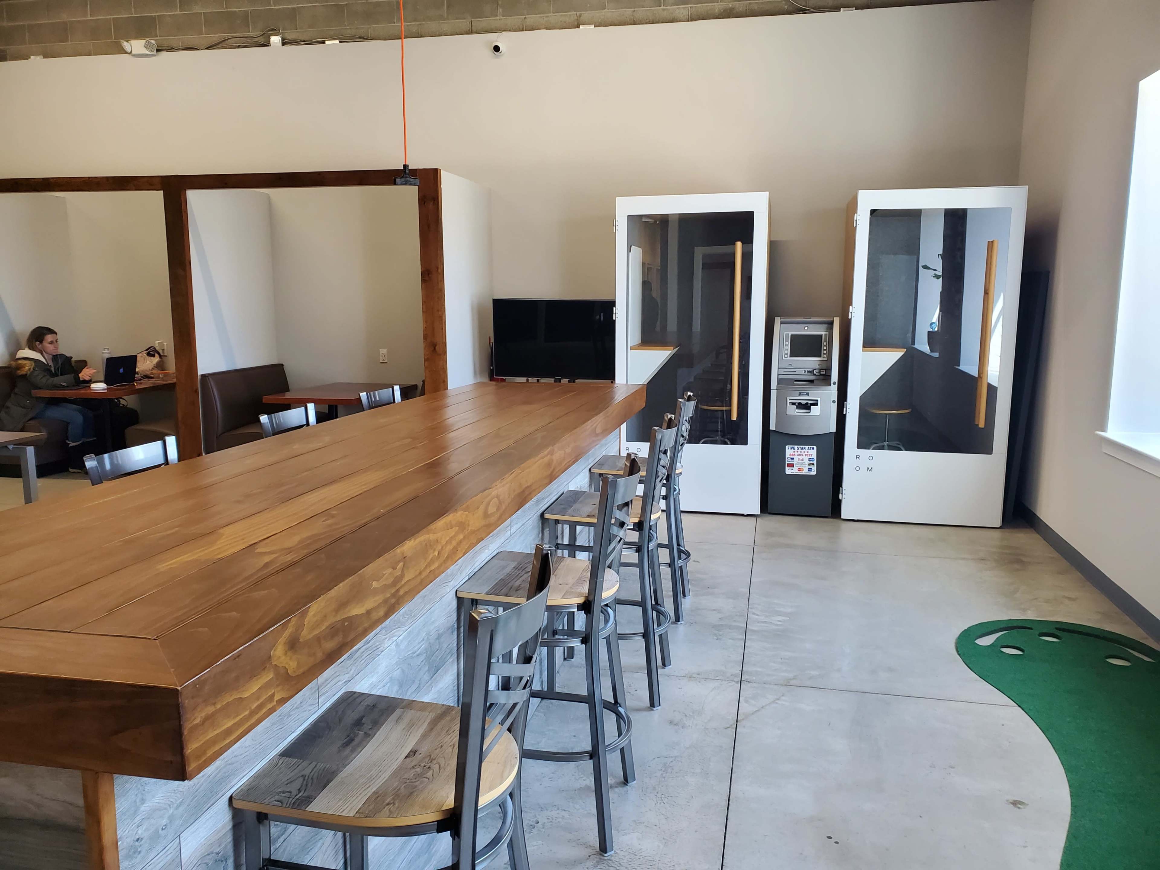 A spacious interior features a wooden bar with high stools, a vending machine, and two white phone booths, with a person working on a laptop in the background.
