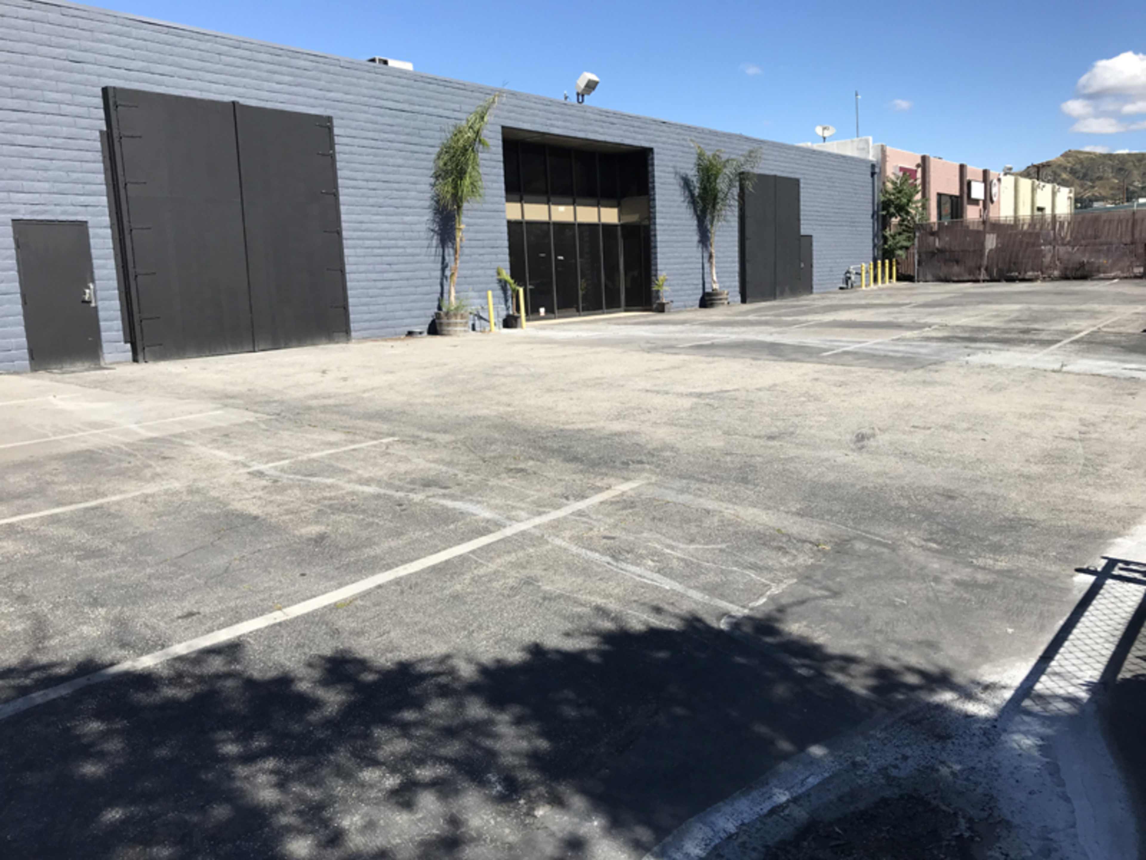 The image shows a large, empty parking lot in front of a gray building with two large black doors and a glass entrance.