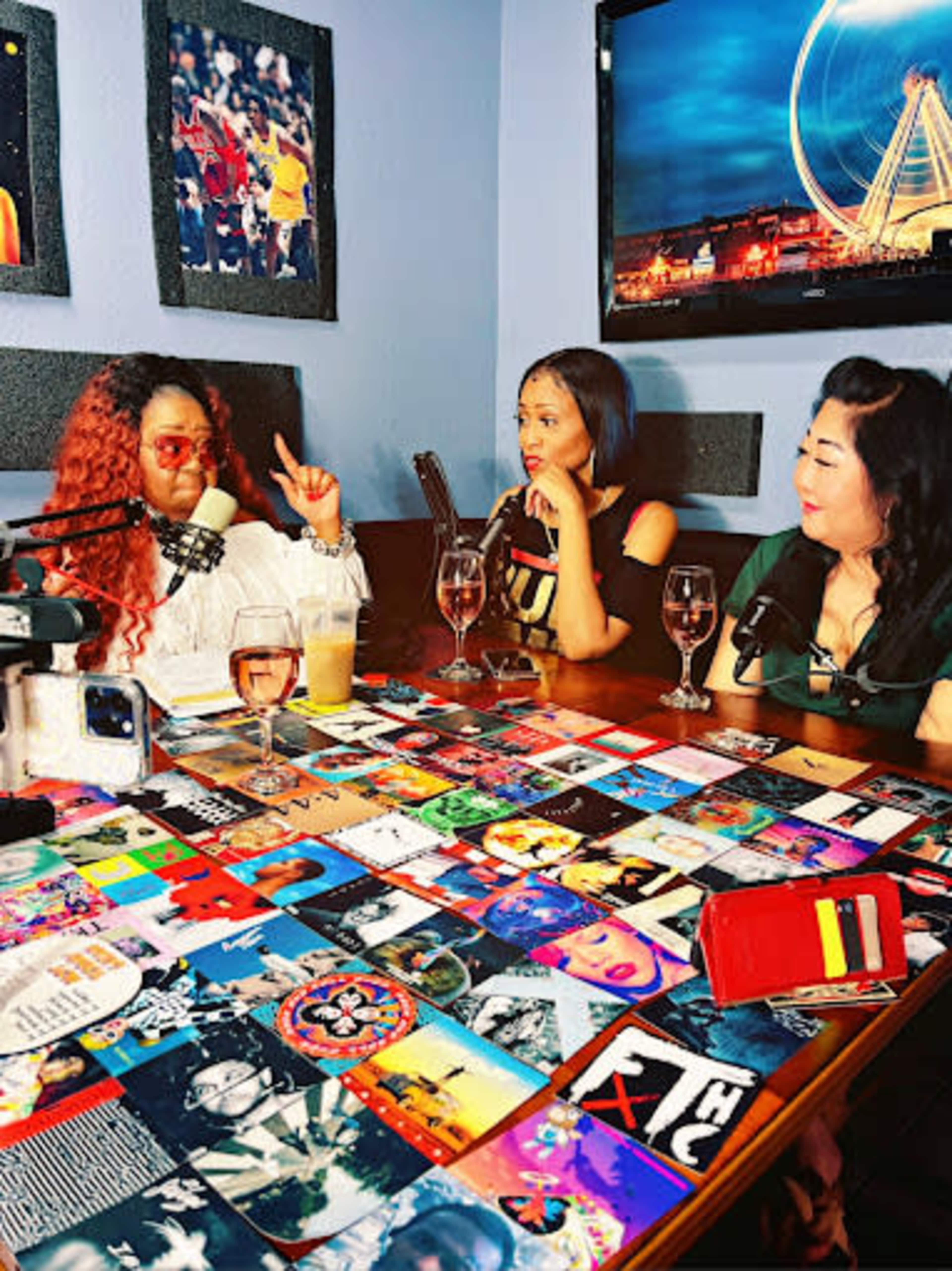 Three women sit at a table covered with colorful coasters, engaged in conversation, with drinks and a camera nearby.