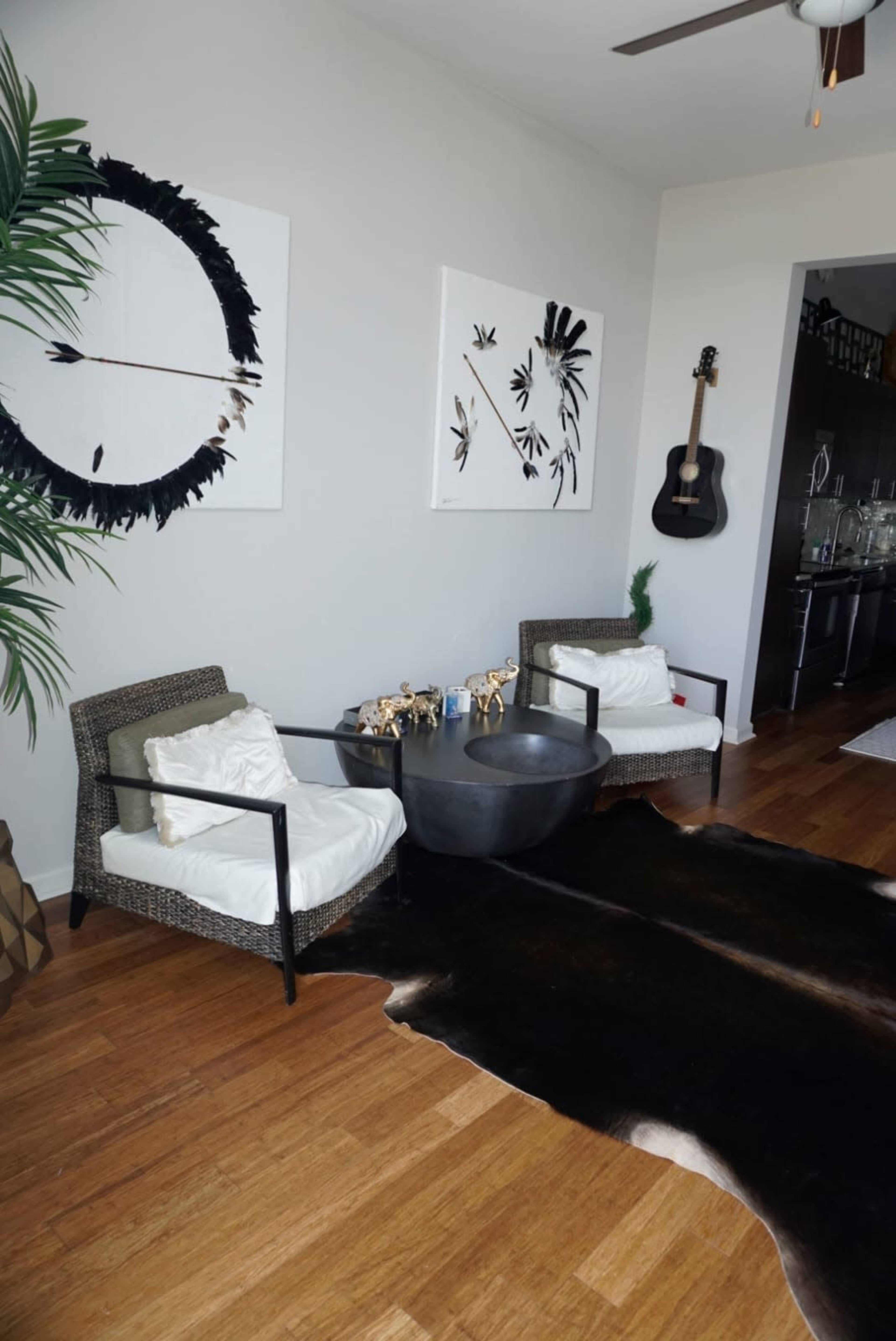 A cozy corner features two wicker chairs with white cushions, a round black table, and a cowhide rug on the wooden floor, flanked by wall art and a guitar.
