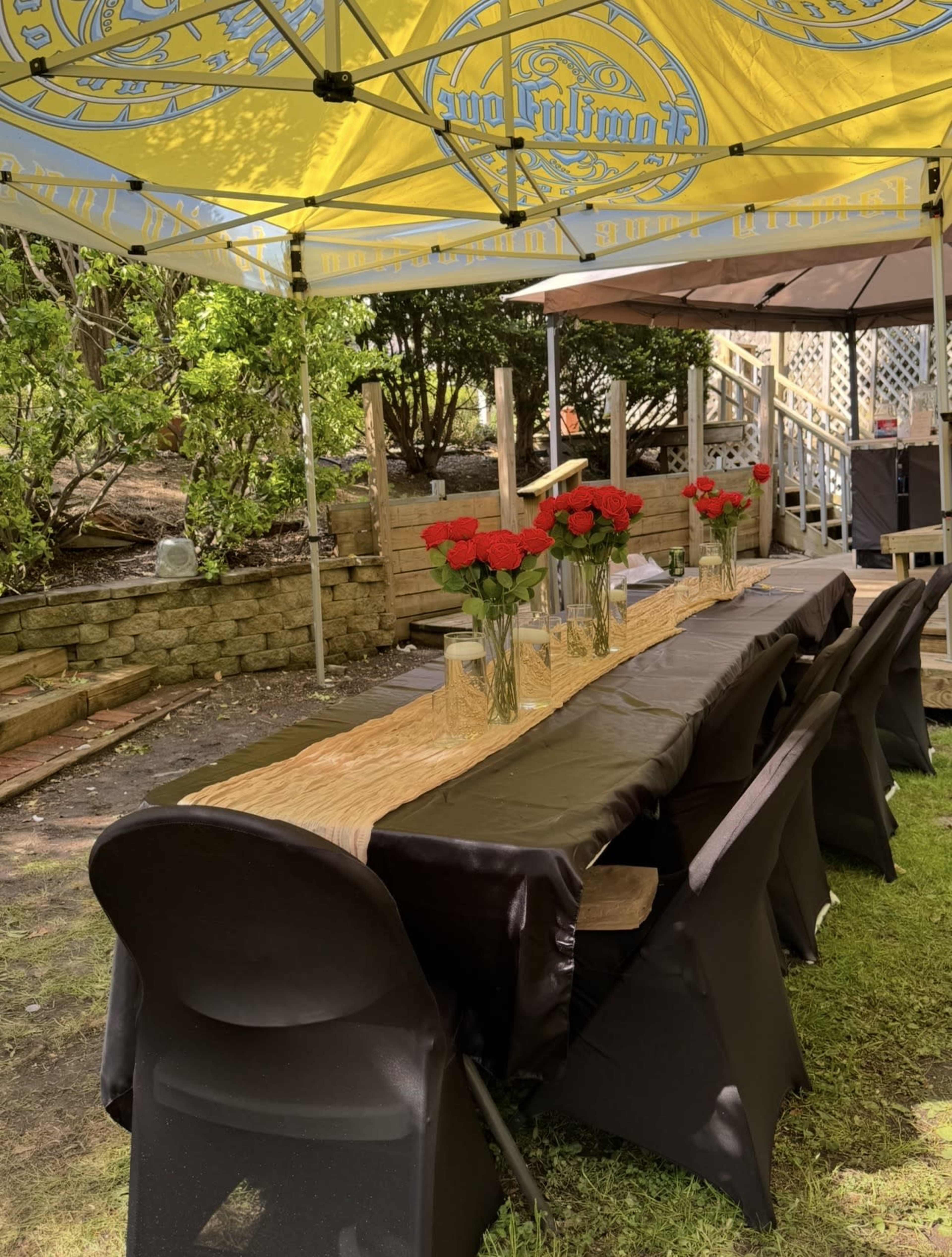A long table is set up under a canopy with yellow and black fabrics, adorned with vases of red roses.