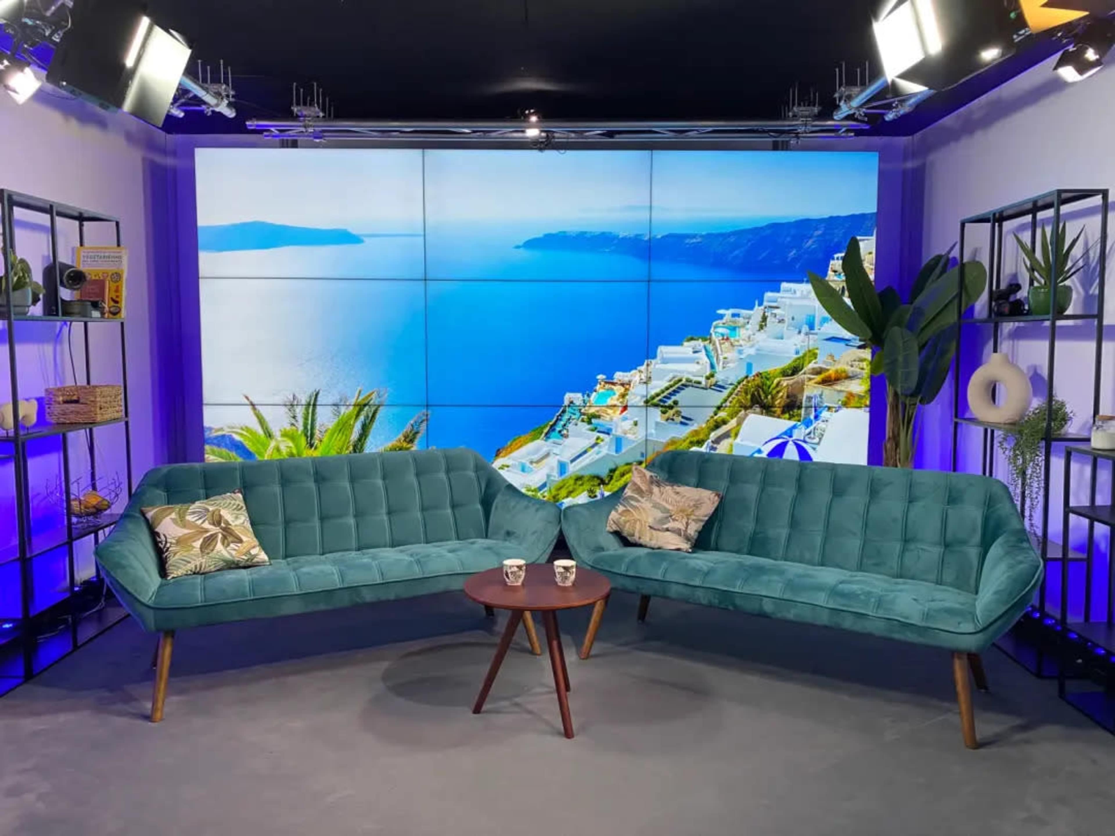 A modern television studio setup featuring two teal sofas and a small round table, with a large screen displaying a coastal view in the background.