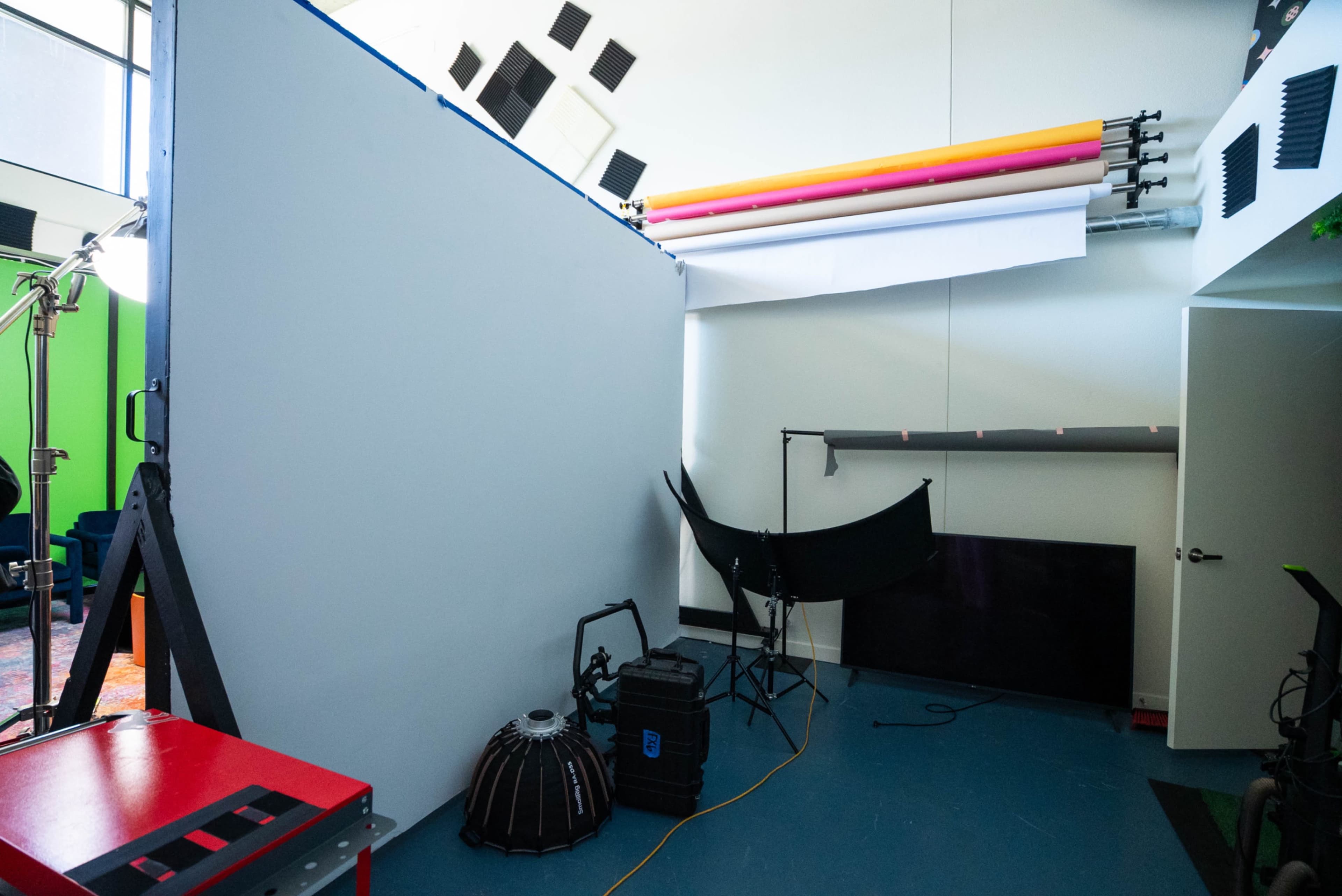 The image shows a partially decorated studio space featuring a blank wall, colorful backdrops, and various photography equipment.