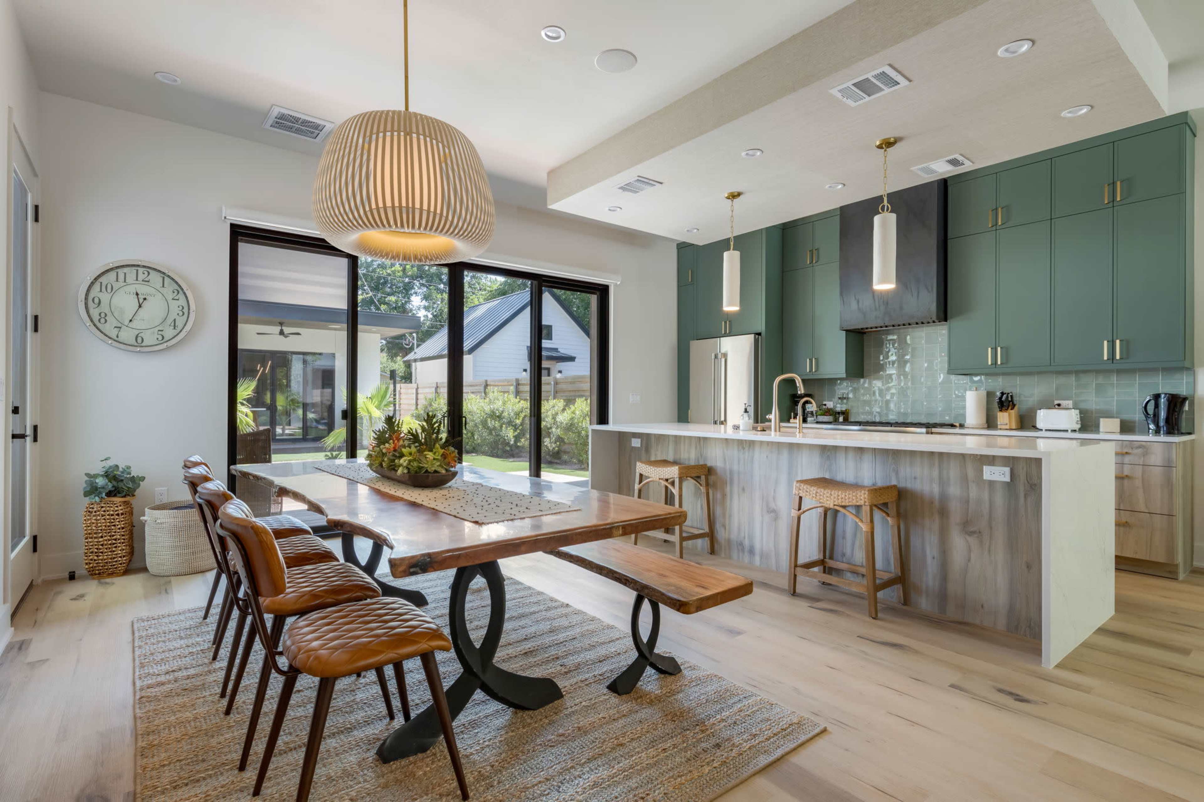 A modern kitchen and dining area features a wooden dining table surrounded by leather chairs, with green cabinets and large glass doors leading to an outdoor space.