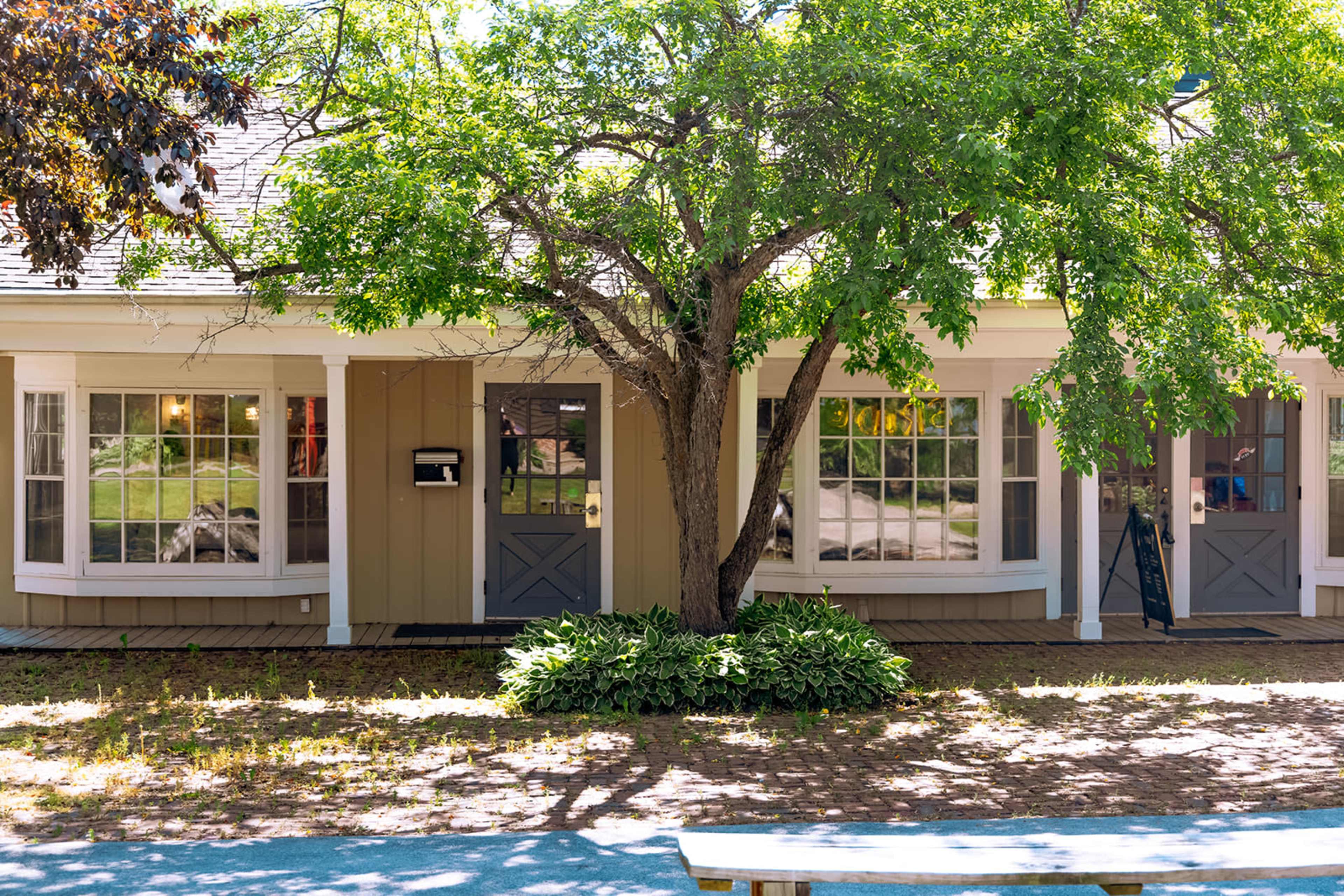 A two-story house with large windows and a central door is framed by a mature tree in front and a wooden walkway leading to it.