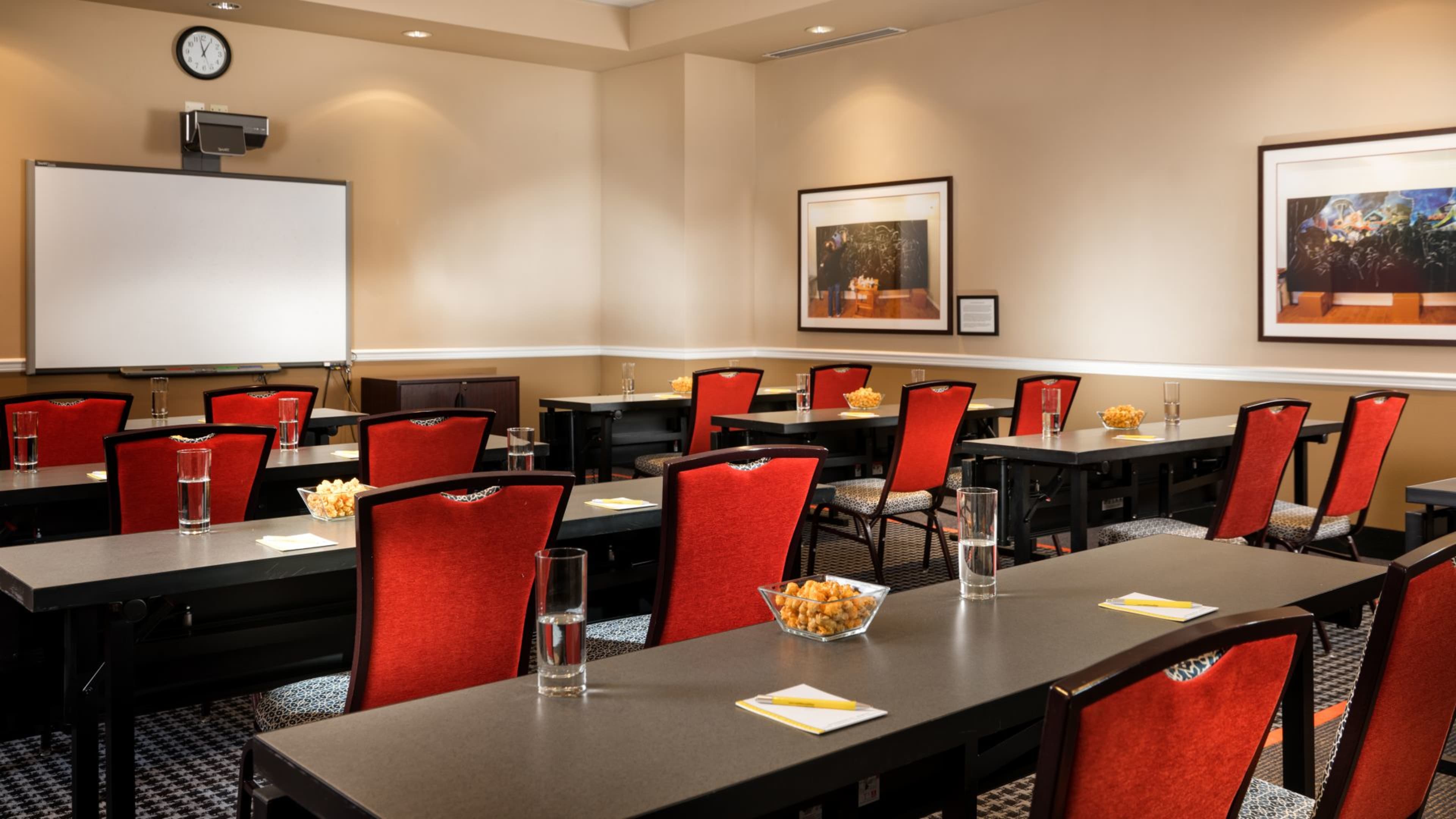 A conference room arranged with rows of tables and red chairs, featuring a whiteboard and framed artwork on the walls.