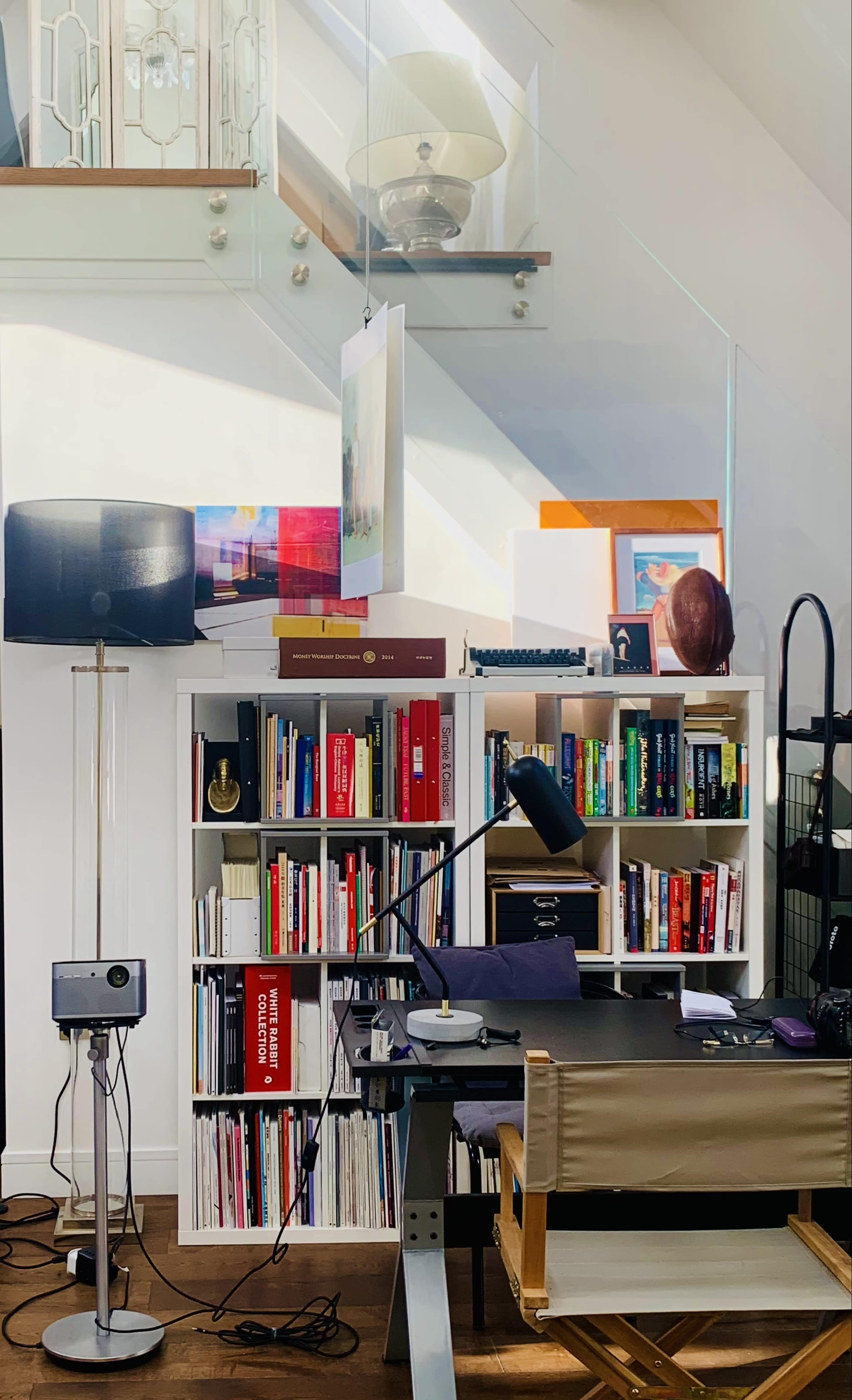A well-organized home office features a bookshelf filled with books, a desk with a lamp and various supplies, and a tall lamp illuminating the space.