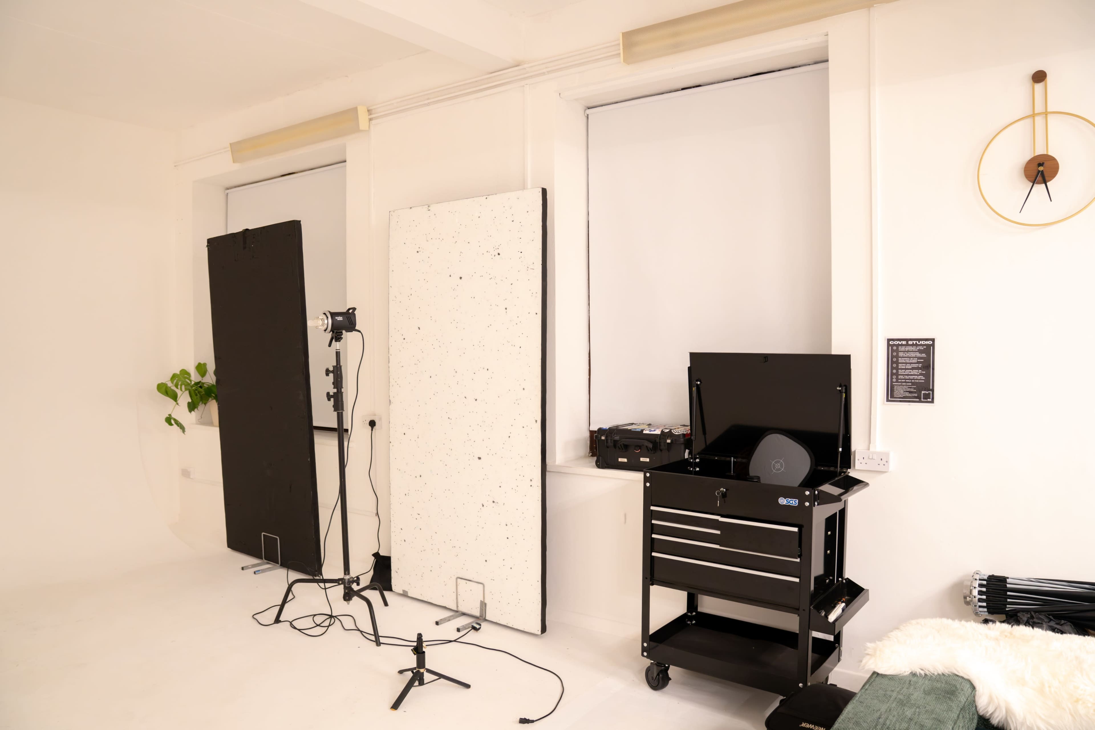 The image shows a minimalist studio room equipped with light stands, a portable backdrop, and a mobile storage cart, all set against a white wall.