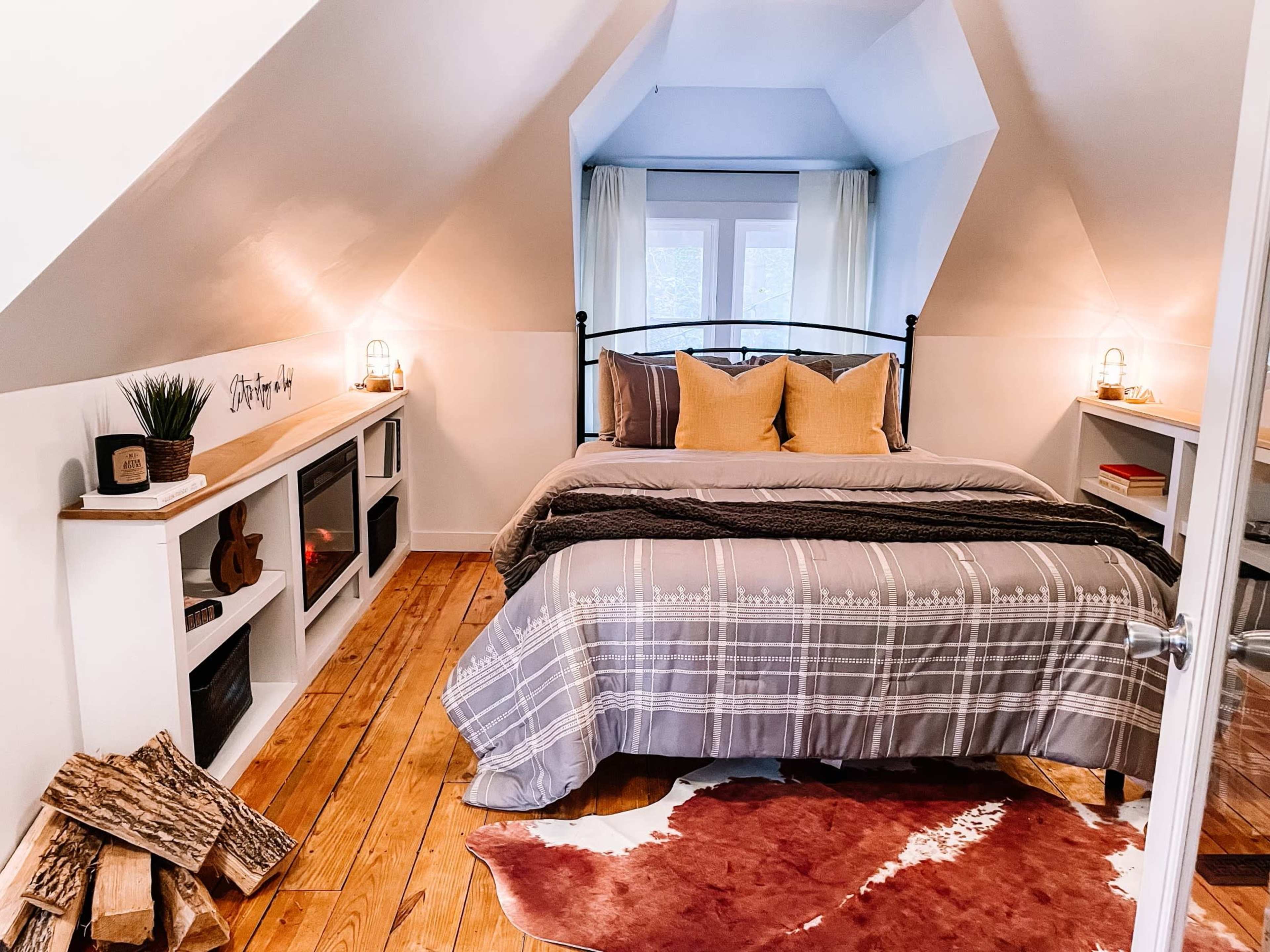 A cozy attic bedroom featuring a bed with gray bedding, decorative pillows, wooden flooring, and rustic accents, including stacks of logs and a cowhide rug.