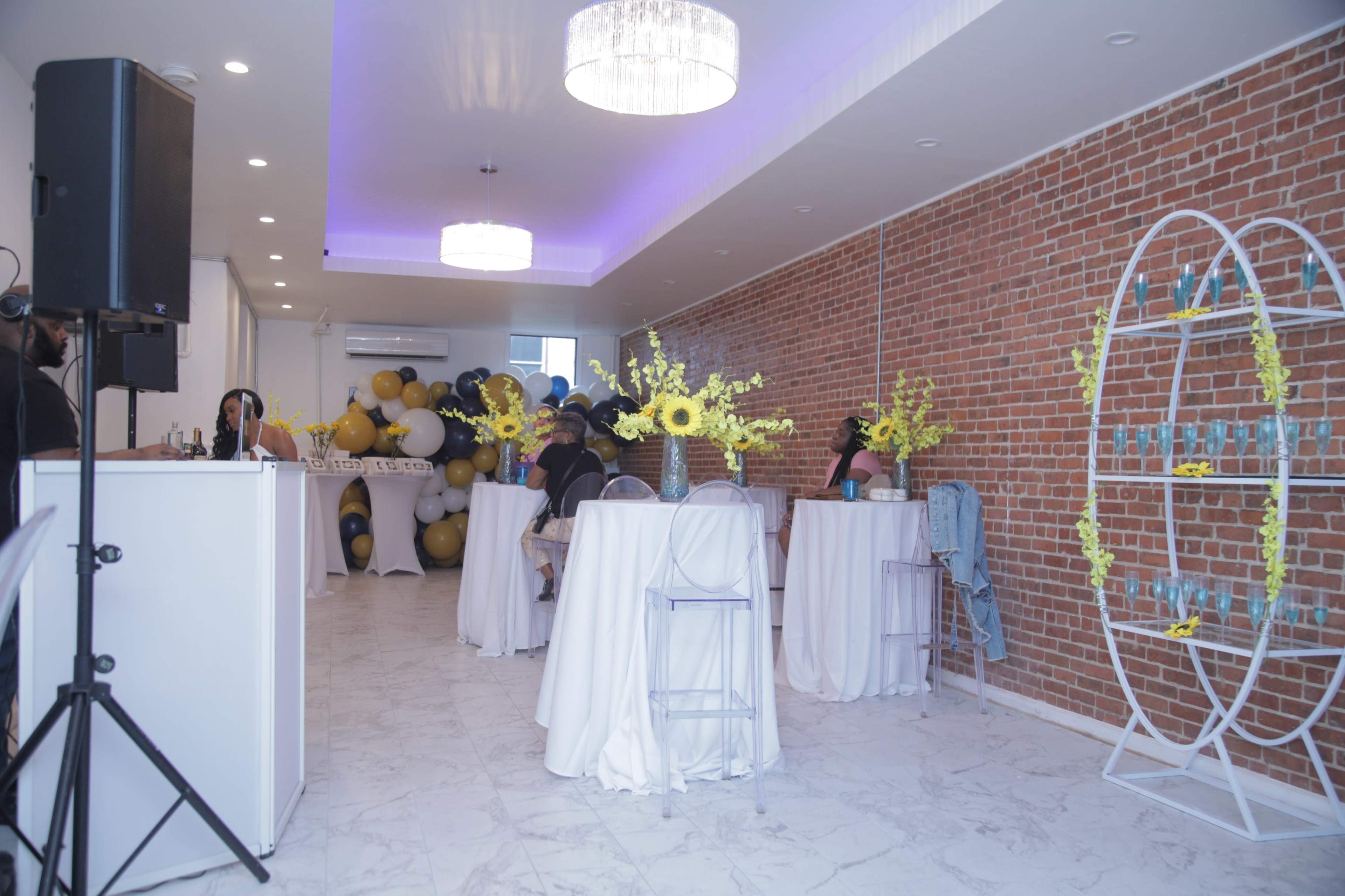The image shows a spacious event room with white tables and chairs, decorated with yellow flowers, a large balloon arrangement, and a drink display.