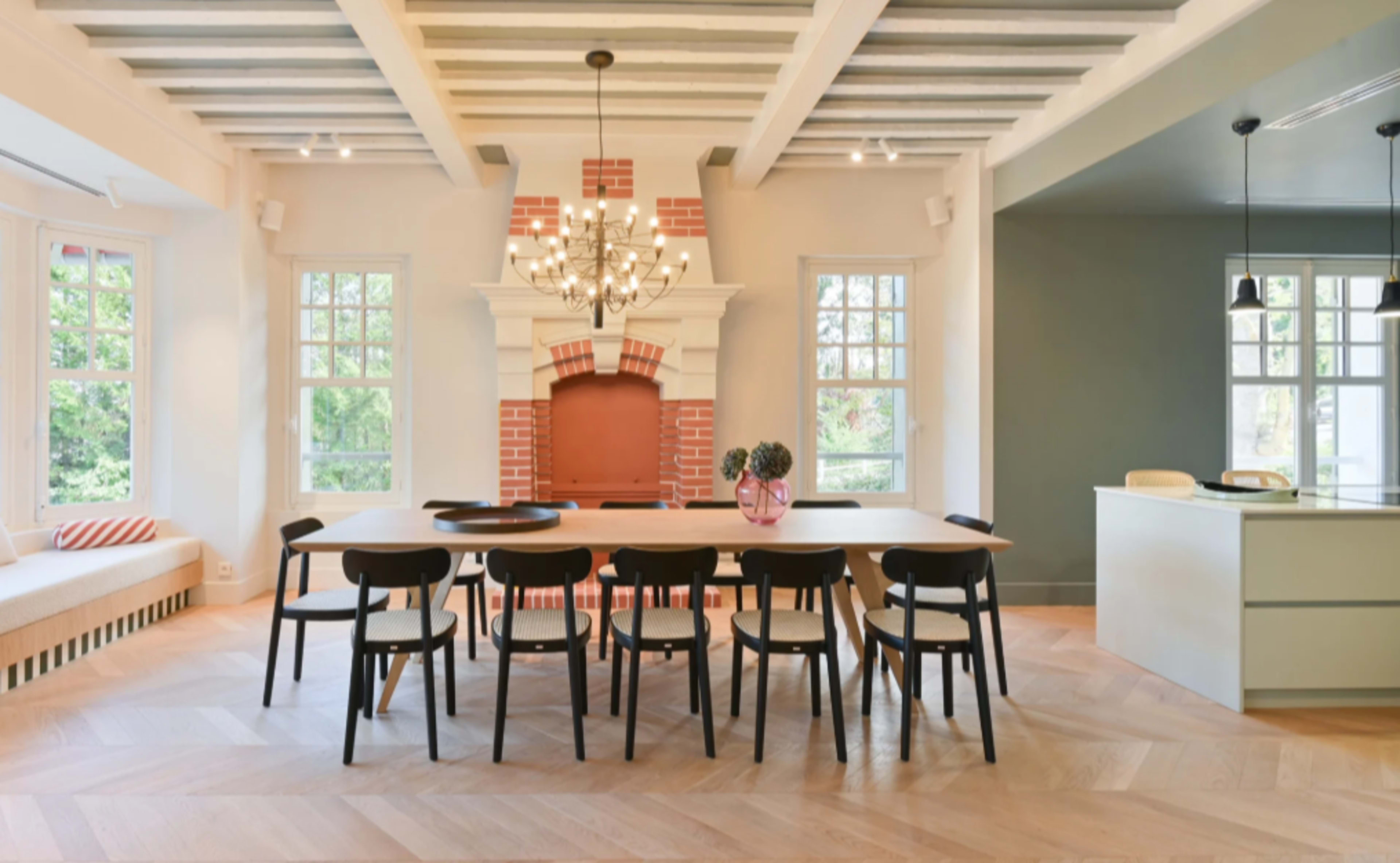The image features a modern dining area with a long table surrounded by black chairs, a large fireplace, and large windows letting in natural light.