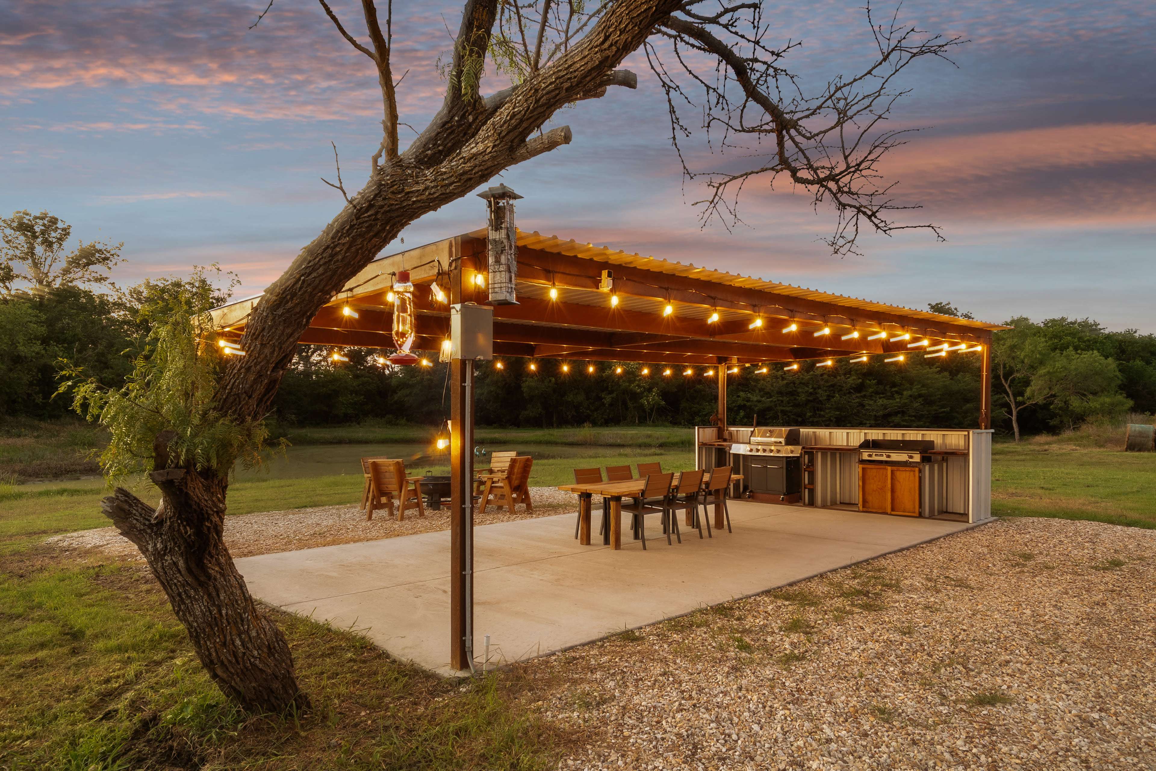 A covered outdoor space features string lights, a grilling area, and a dining table set up on a gravel surface.