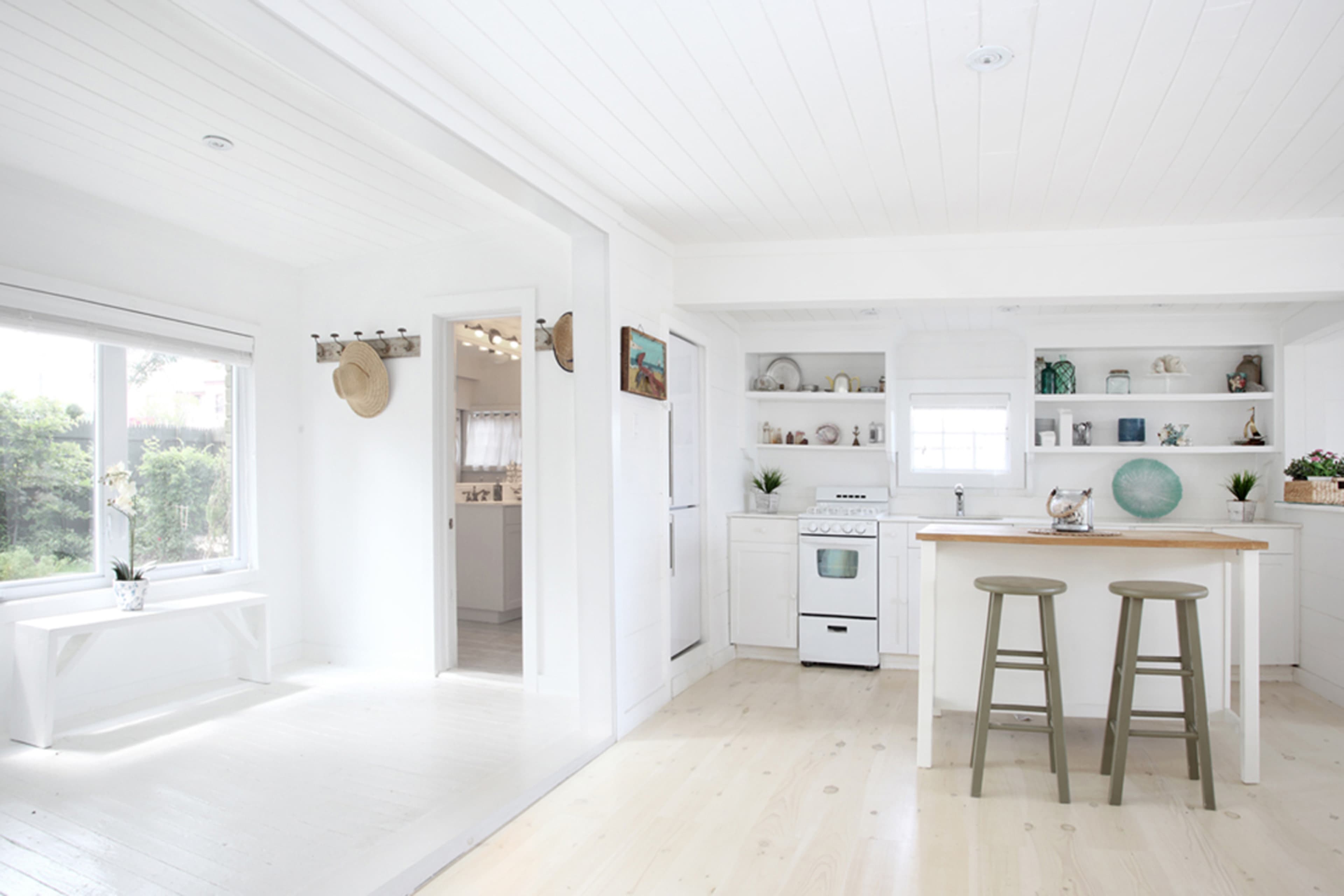 A bright, open kitchen and living area with white walls, wooden floors, and a small island with two stools.