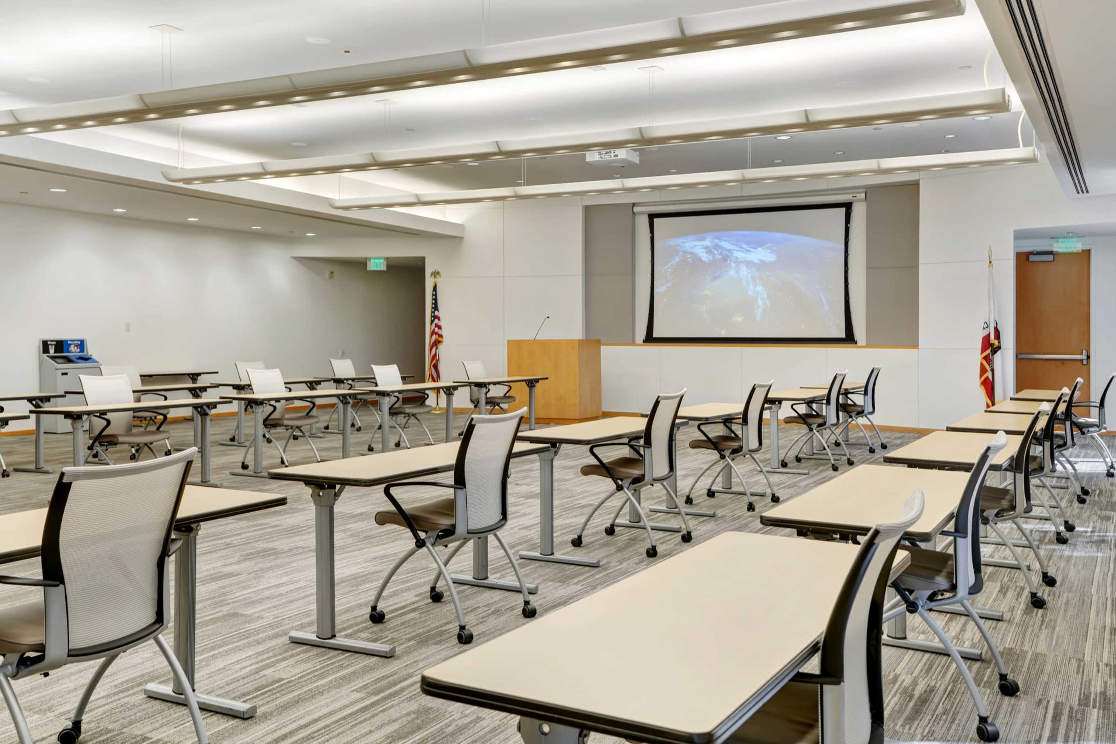 The image shows a modern classroom setup with several movable desks arranged in a spacious layout, featuring a projector screen displaying a global map at the front.