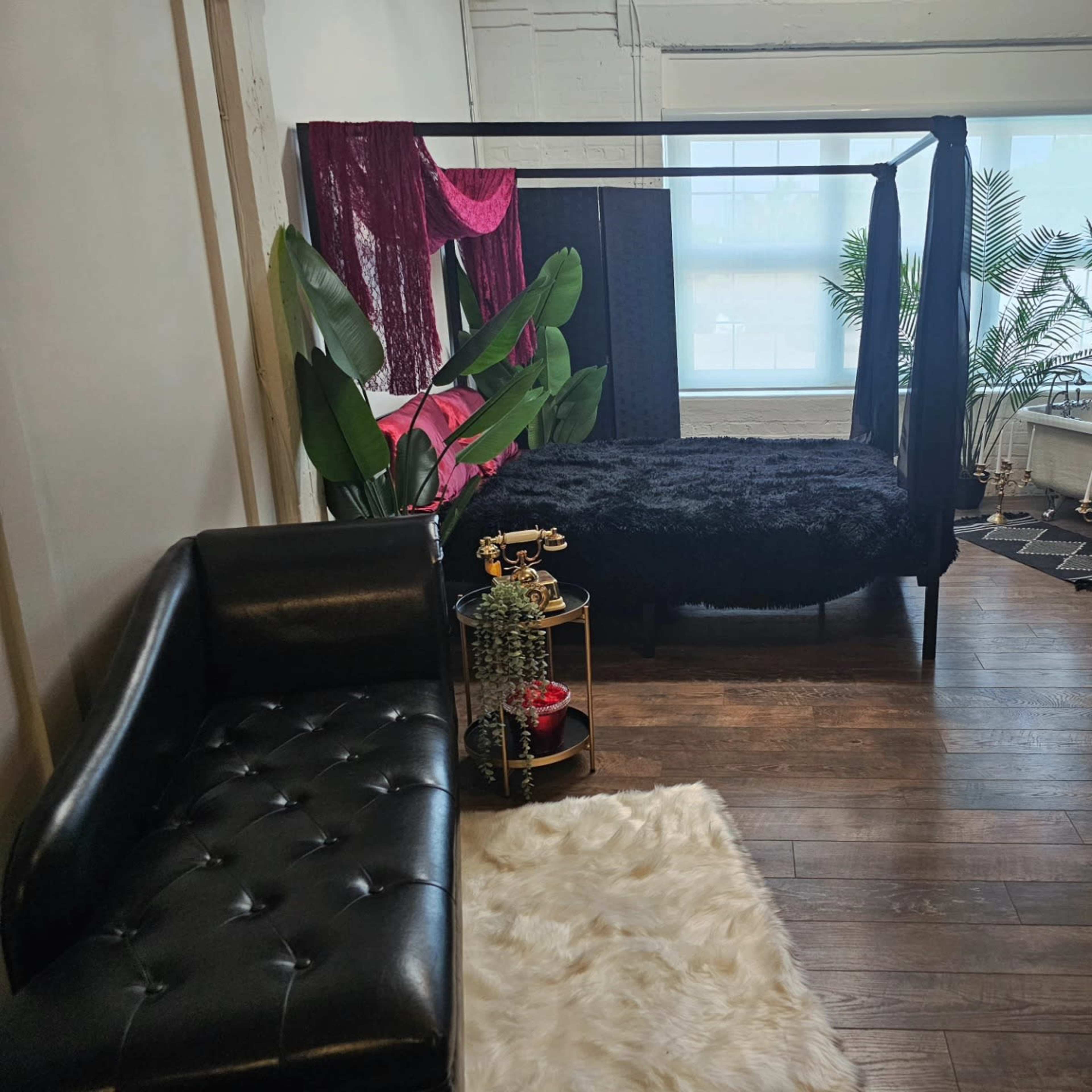 The room features a black canopy bed adorned with textiles, a black leather couch, and a small table, all set against a backdrop of plants and natural light.