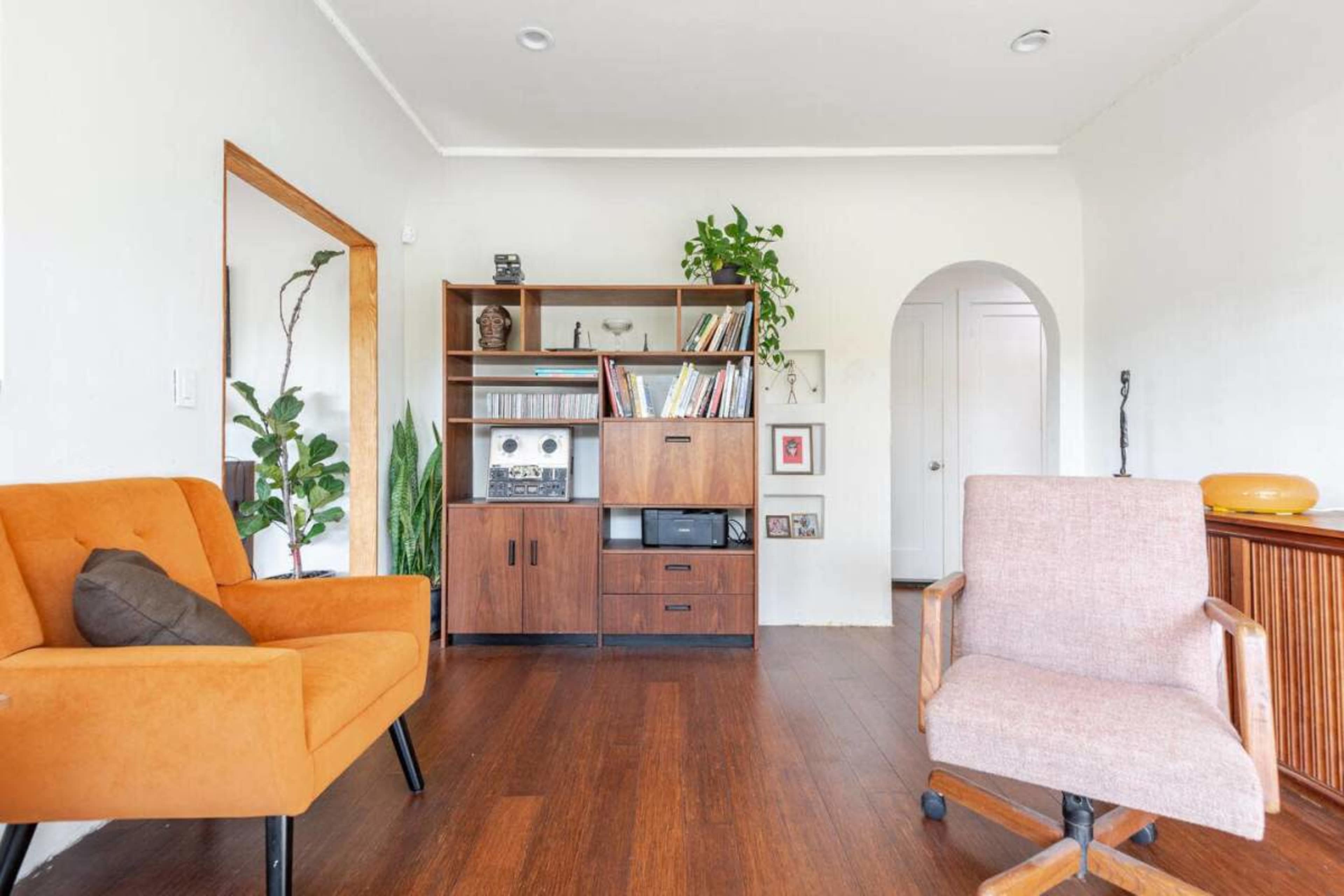 The image shows a modern living room with two armchairs, a bookshelf, and various indoor plants against a light-colored wall.
