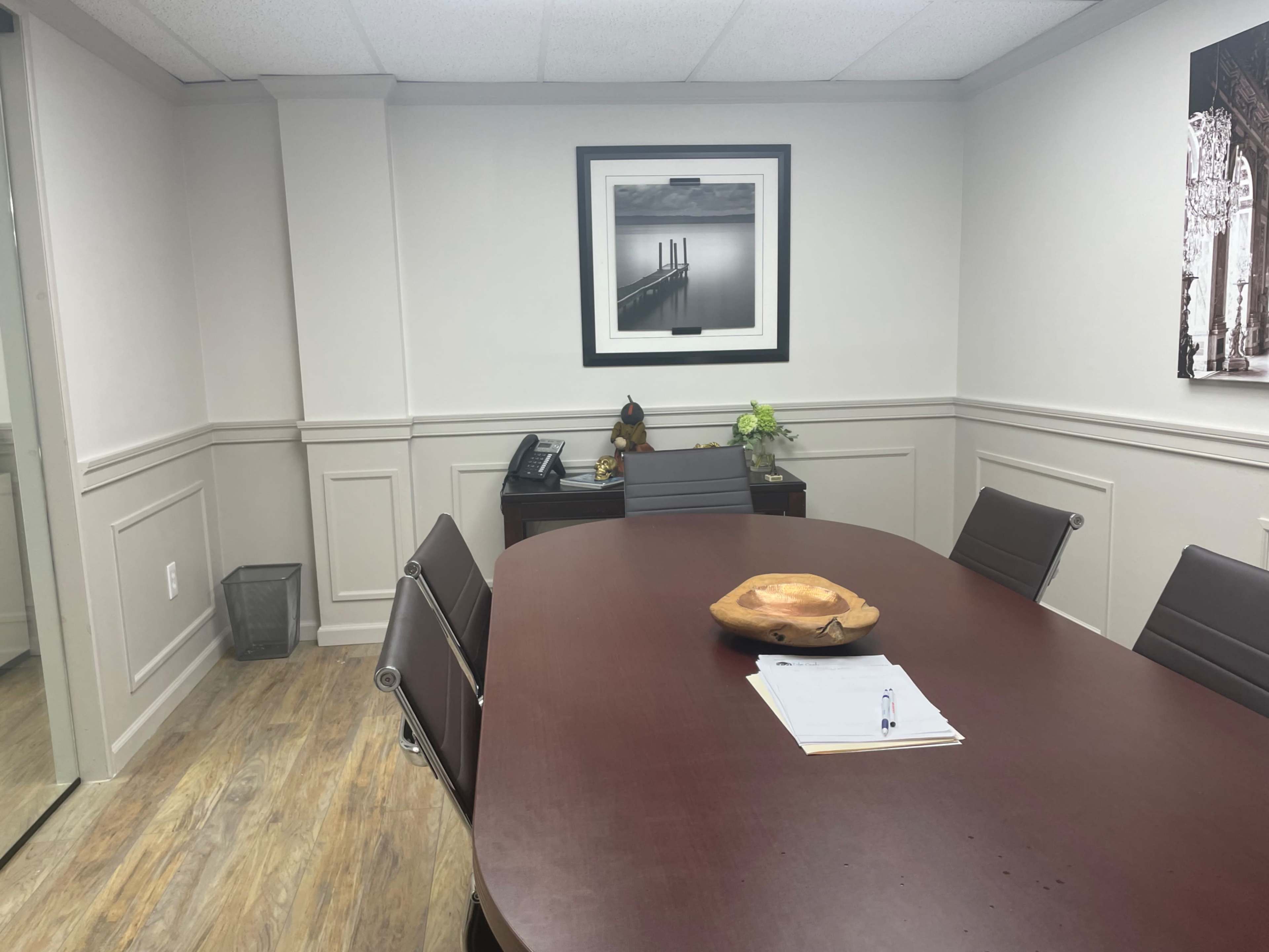A conference room features a long wooden table surrounded by chairs, a notepad on the table, and framed artwork on the walls.