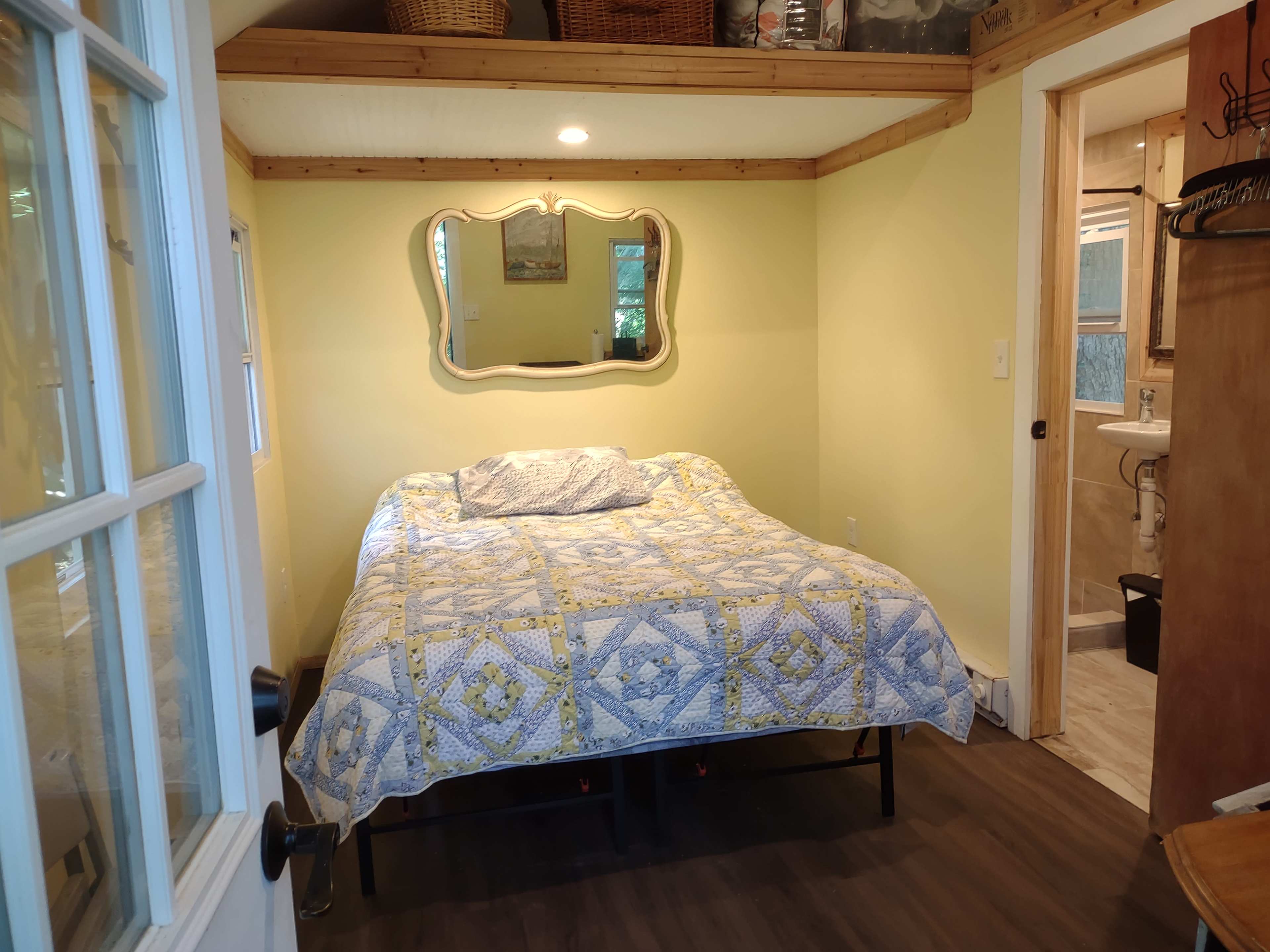 A small bedroom with a bed covered in a patterned quilt, a mirror hanging on the wall, and a doorway leading to a bathroom.