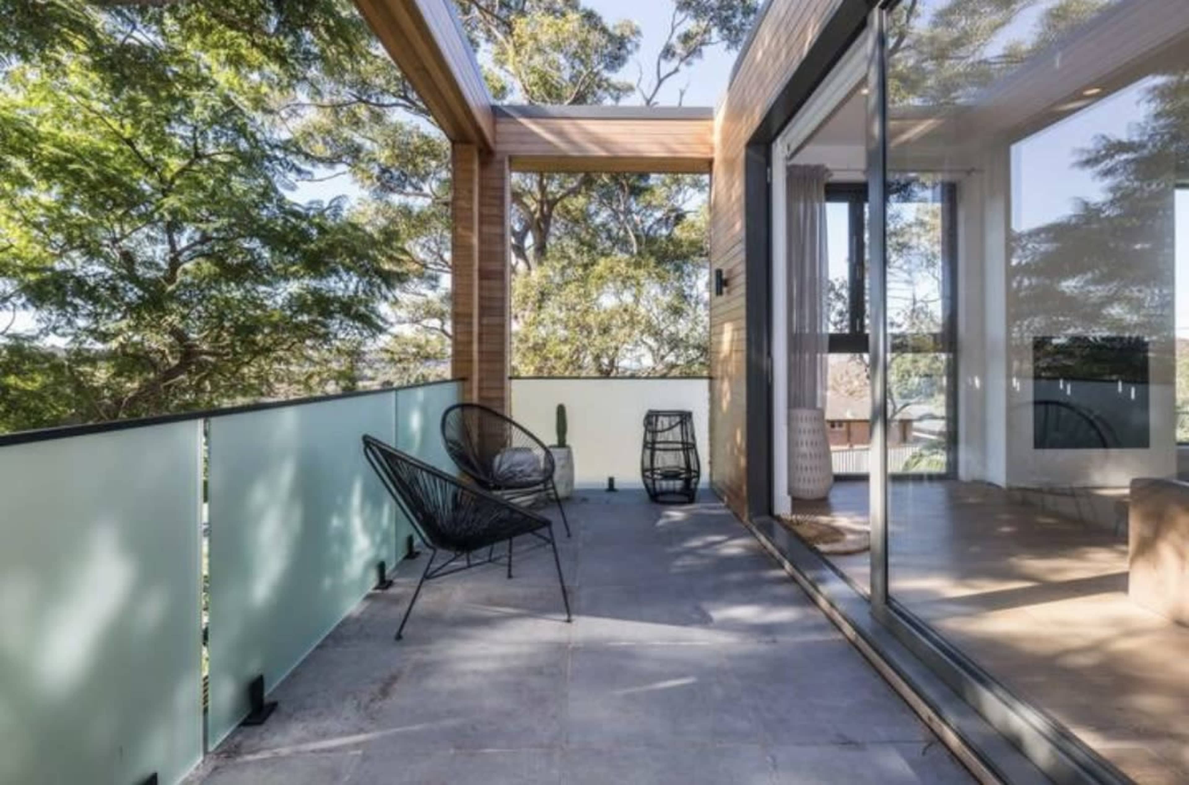 The image shows a spacious balcony with a wooden overhang, a concrete floor, and large glass doors leading into a modern interior, surrounded by trees.