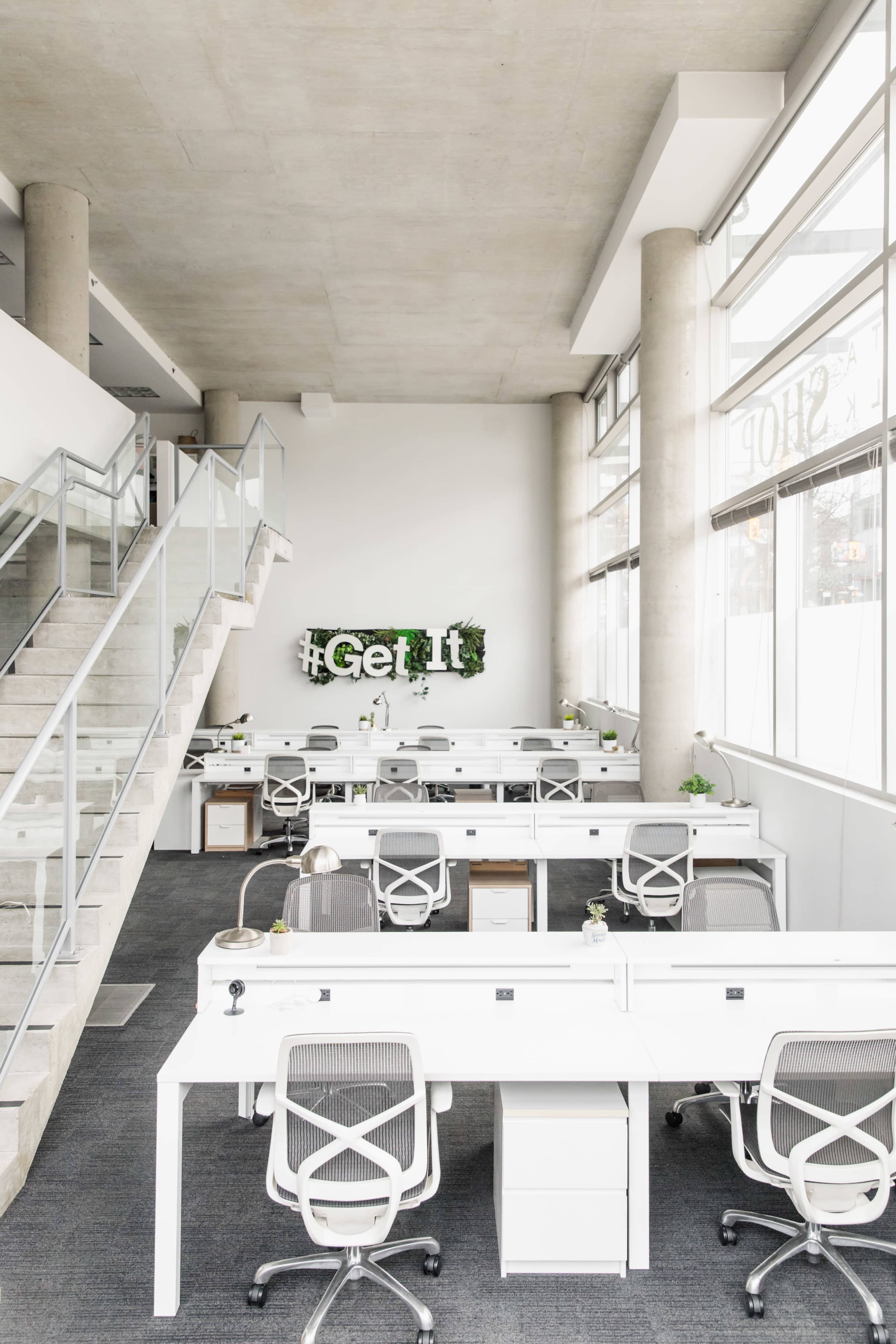 An open office space with white desks and chairs, large windows allowing natural light, and a green wall with the hashtag "#GetIt" on it.