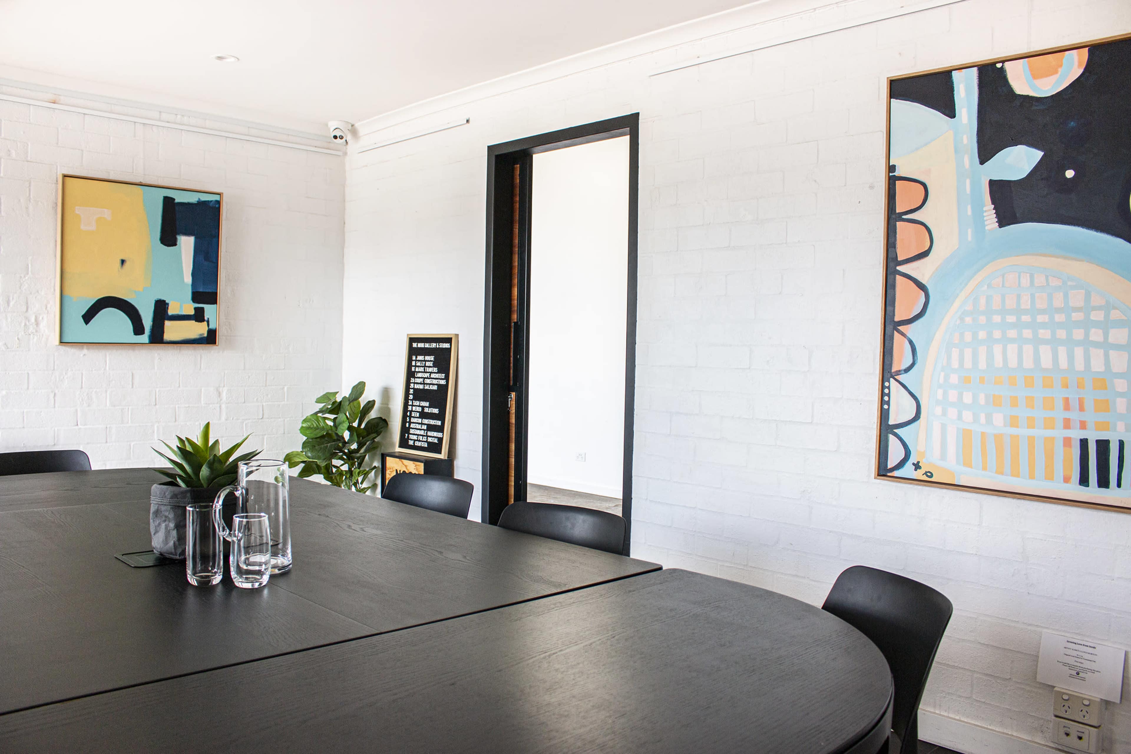 A modern meeting room features a large black table surrounded by chairs and adorned with abstract artworks on white brick walls.