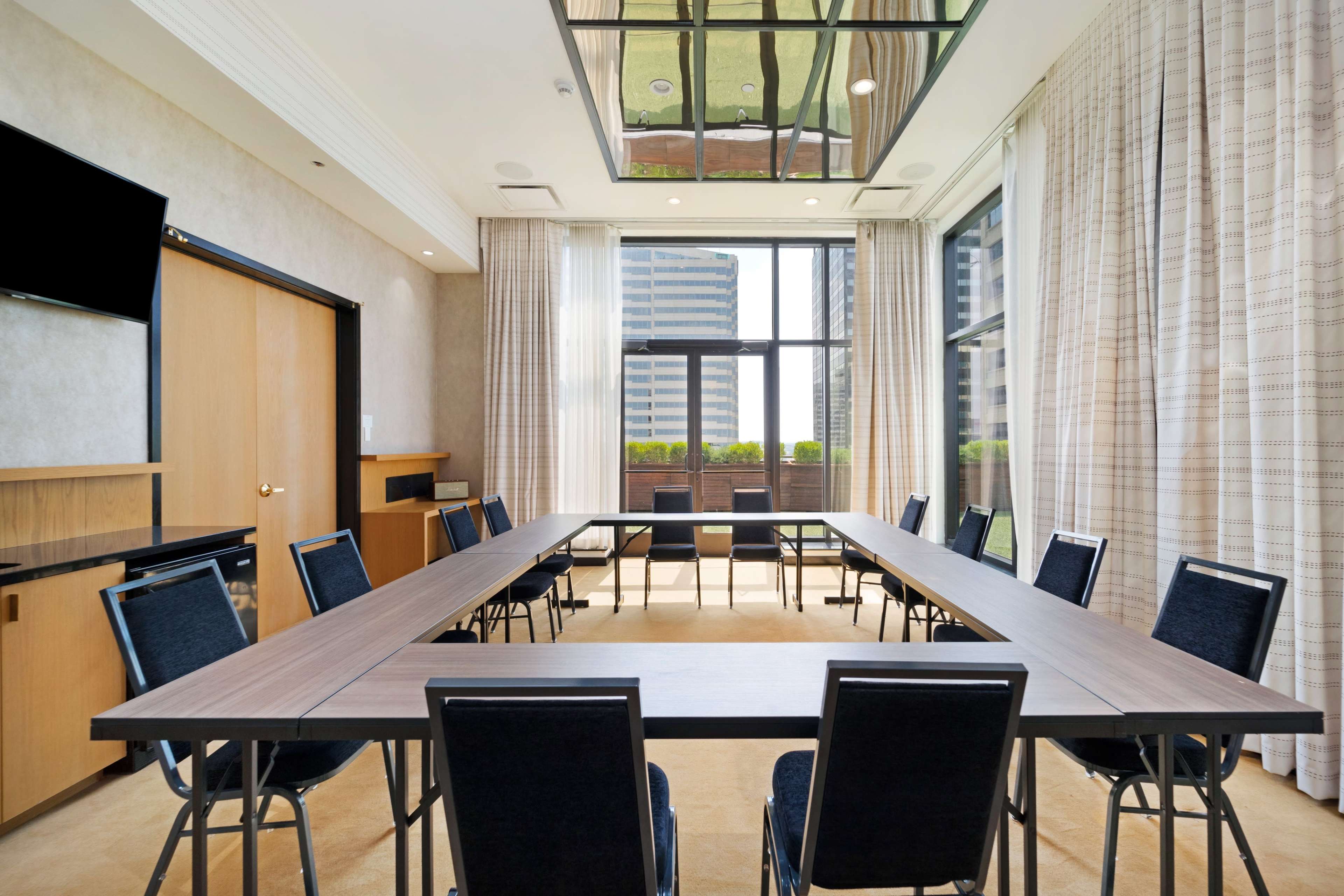 A modern conference room features a large table surrounded by chairs, with large windows letting in natural light and a view of nearby buildings.